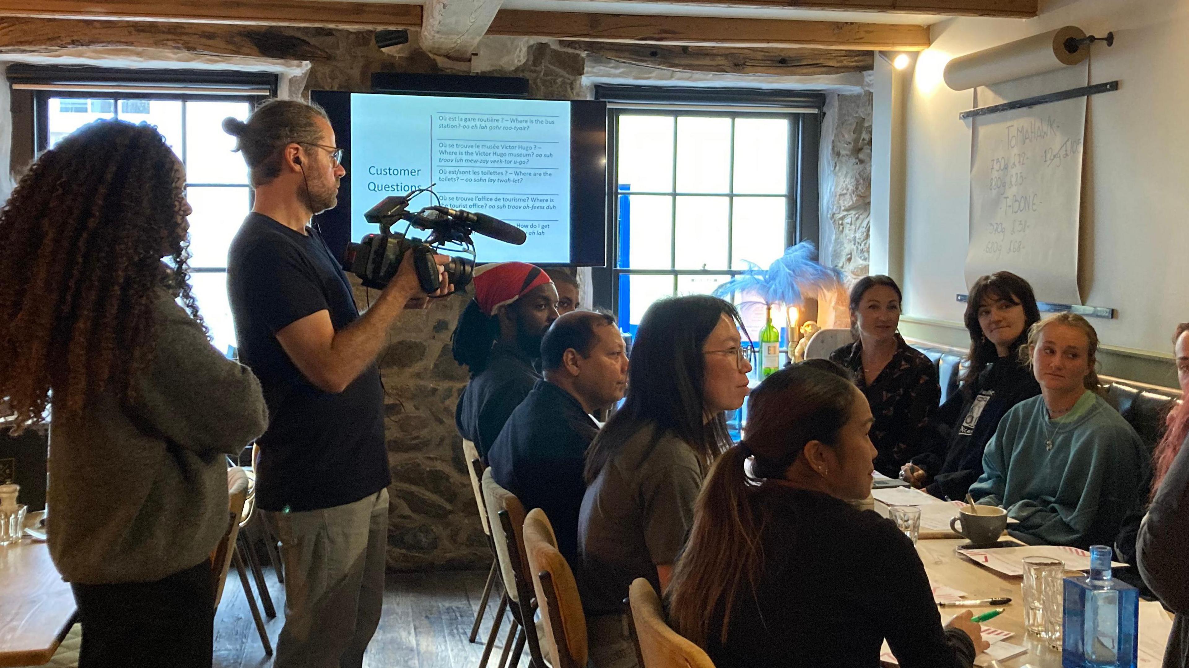 A woman with long curly hair and grey jumper standing next to a man with glasses and a blue t-shirt holding a camera. They are filming a class of people sat around a long table.