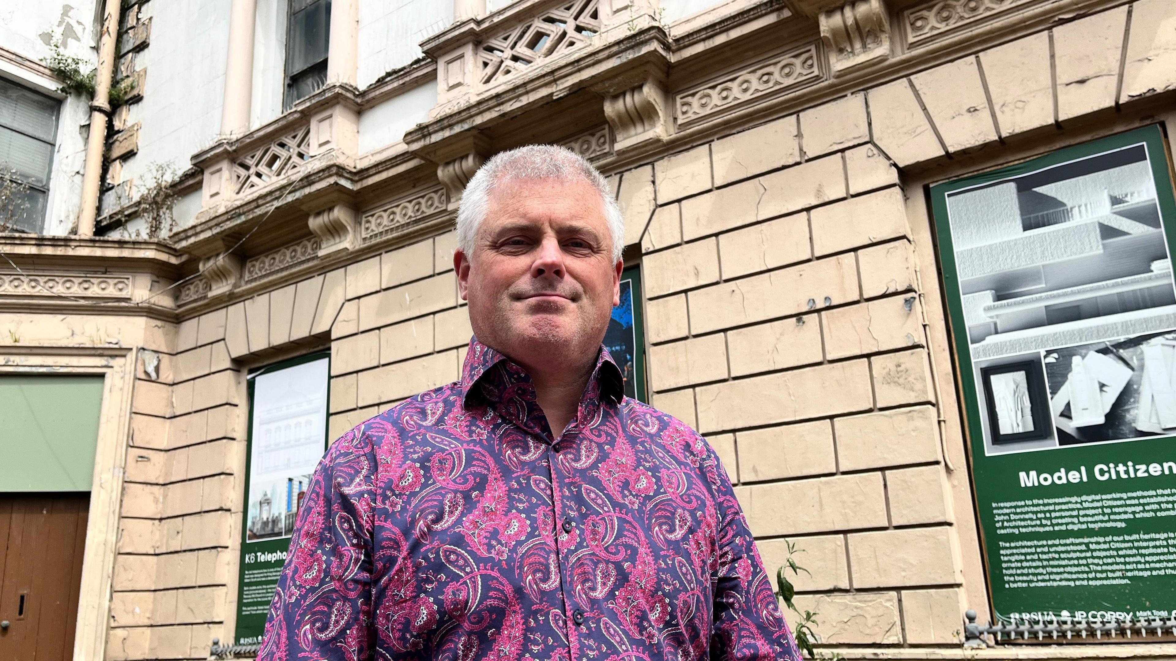 A man stands in front of the beige assembly buildings. He wears a pink and purple shirt. The man has short grey hair. There is a green notice board in place of a window over his left shoulder.