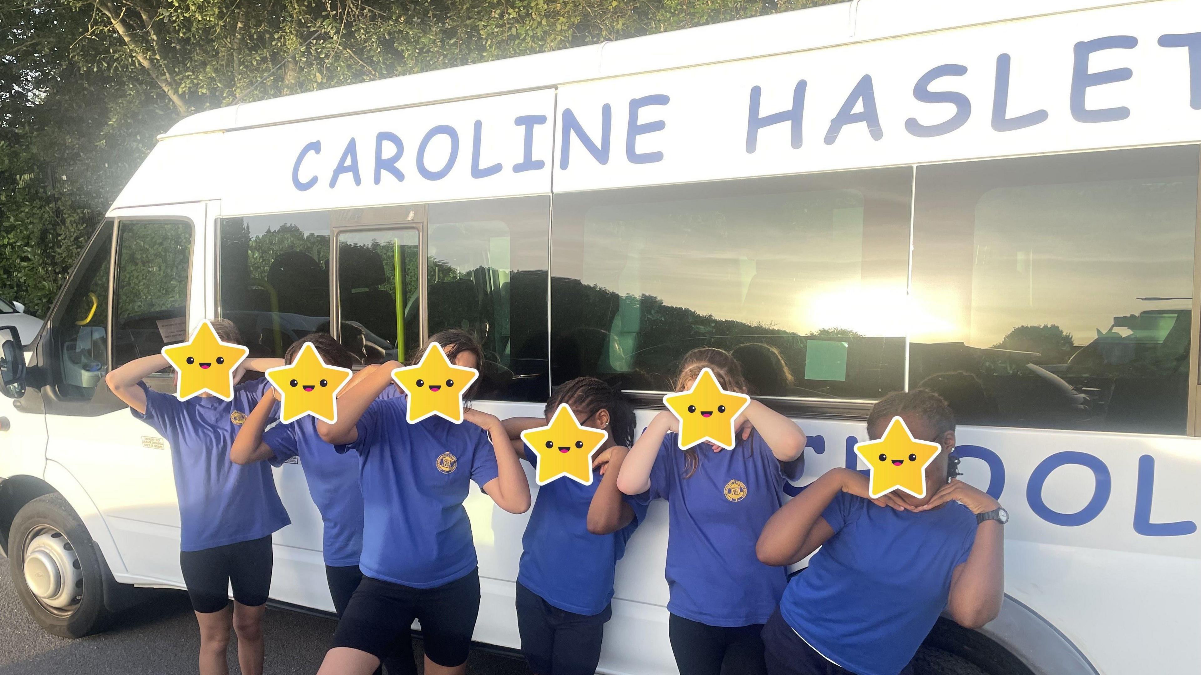 Six pupils from a school standing outside a white school minibus, they all have stars on their faces to hide their identity. They are all wearing blue school tops, with a logo on the right and black shorts. They are posing for an image. 