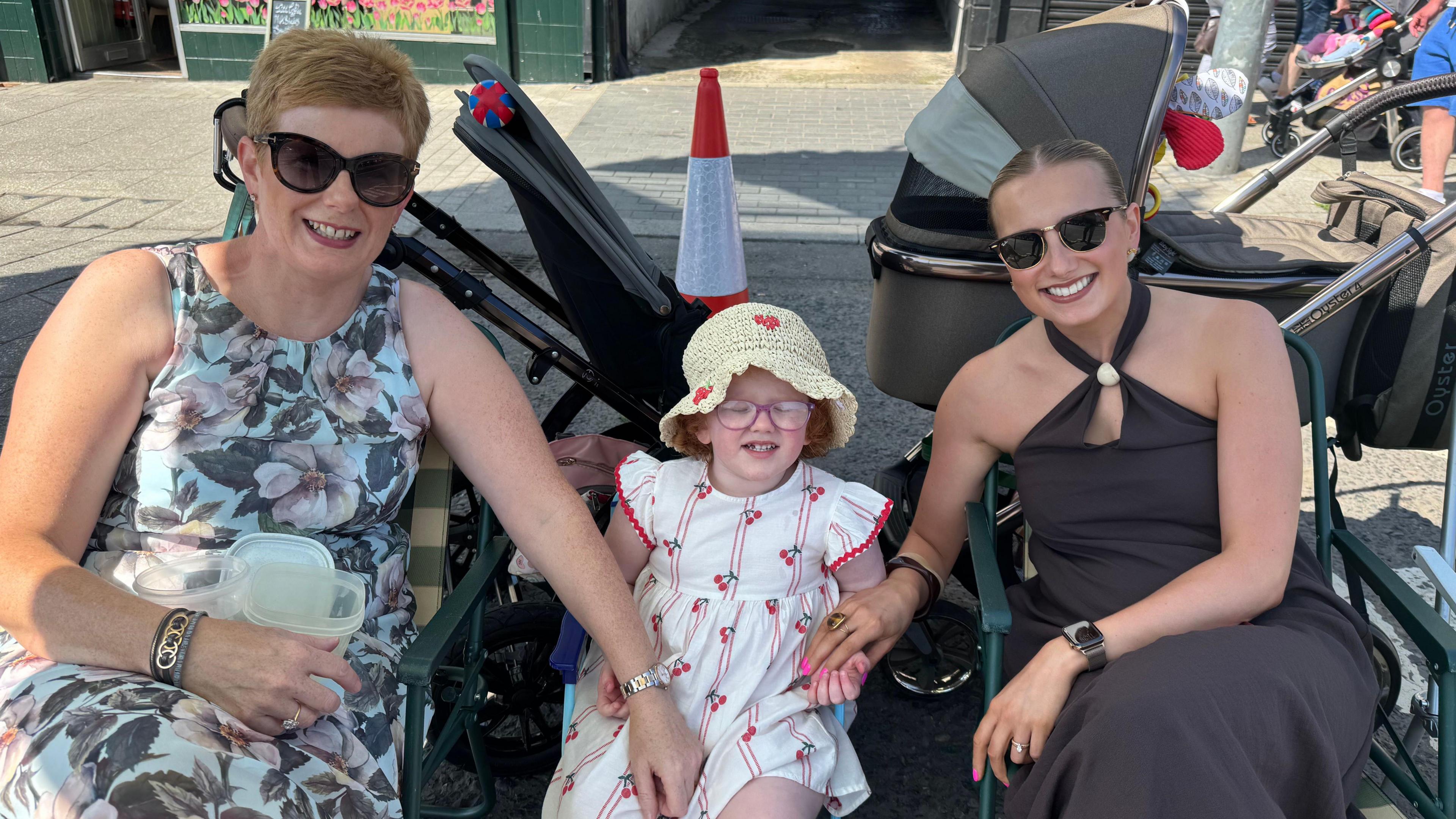 Ellen Watson has light brown hair tied back an parted in the middle and is wearing sunglasses. A middle aged woman with short reddish hair and sunglasses is on the left and a young girl with glasses and a sun hat is in the middle