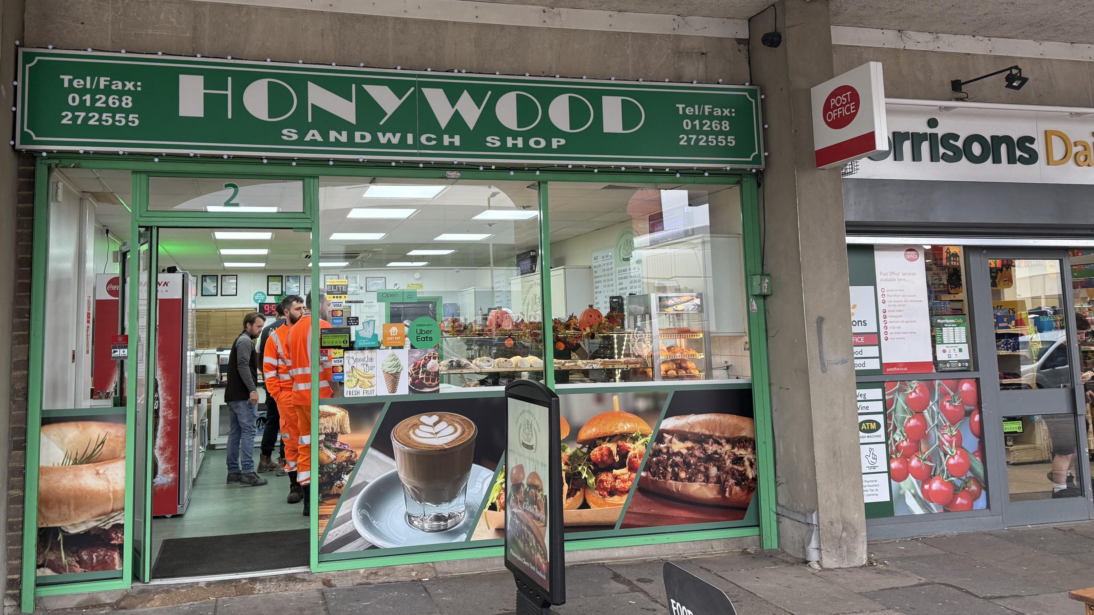 The exterior of a sandwich shop. Above the door is a green sign with the business name - Honywood Sandwich Shop - in white lettering. About five people can be seen inside the shop.