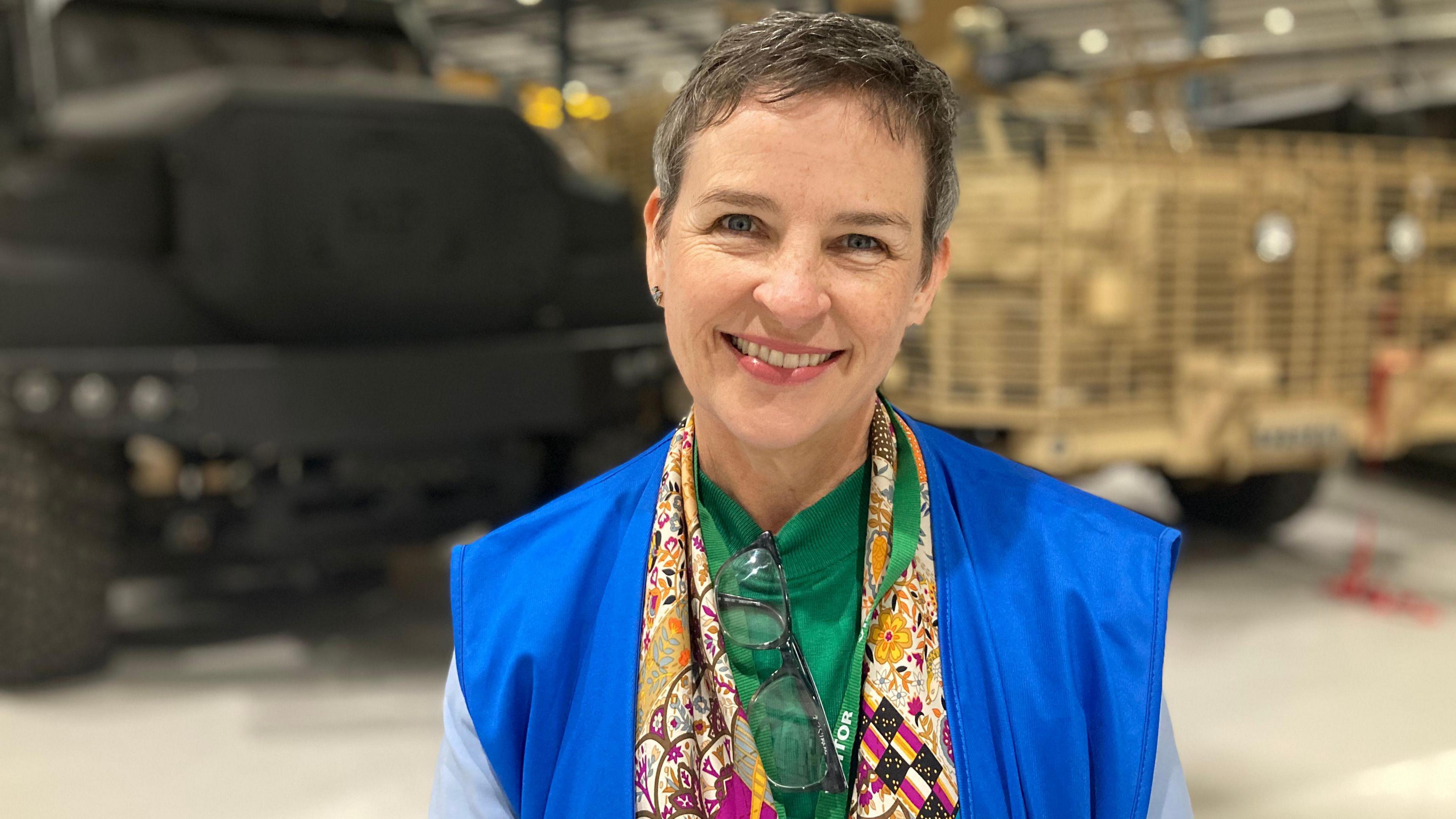 A woman wearing a green top and multicoloured scarf smiles into the camera. Behind her are the blurred outlines of a grey and khaki vehicle.