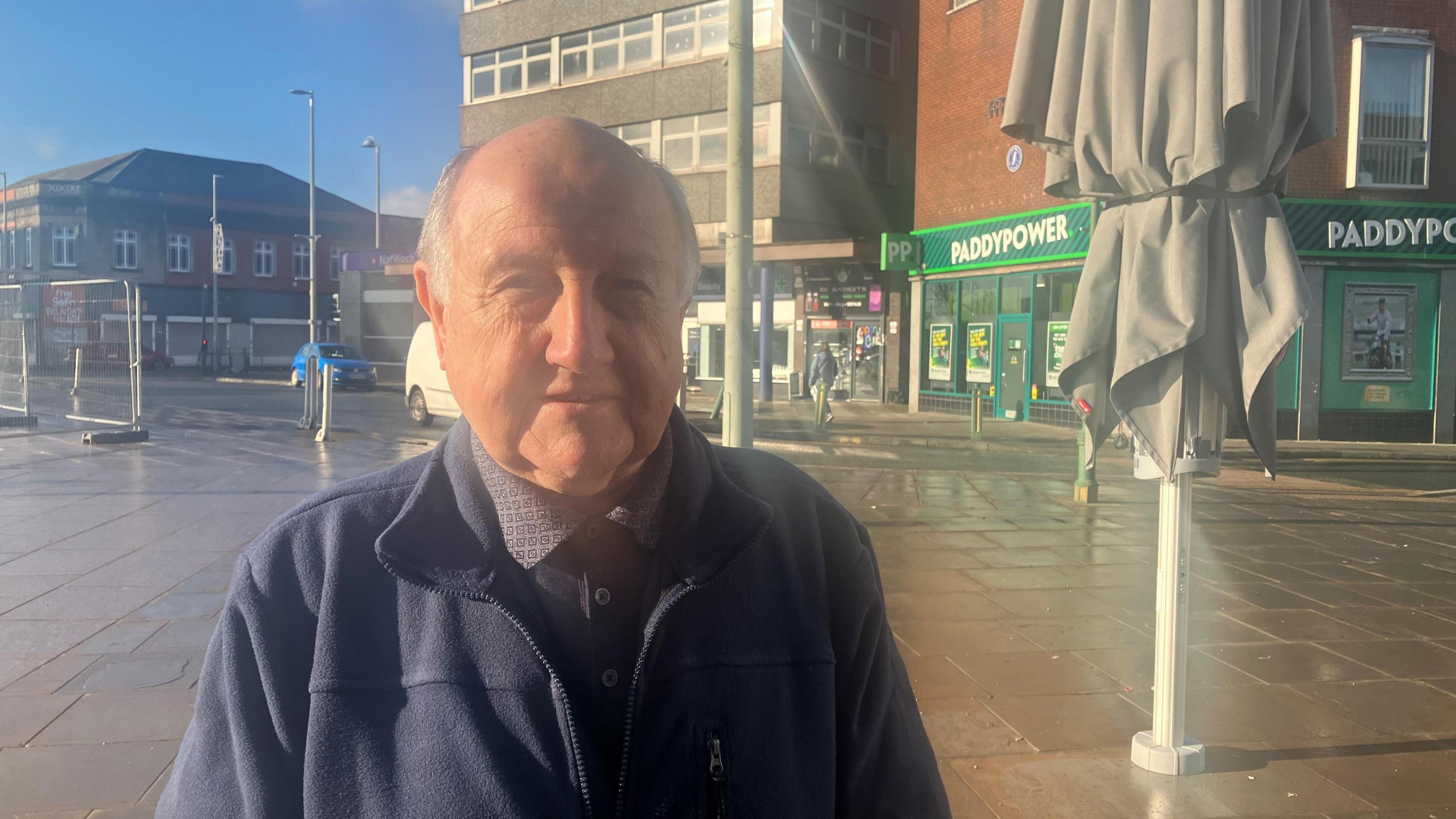 Shopper Steve Hodgson, who is wearing a blue jacket and shirt, stands close to a grey parasol on a sunny day in a town centre. 