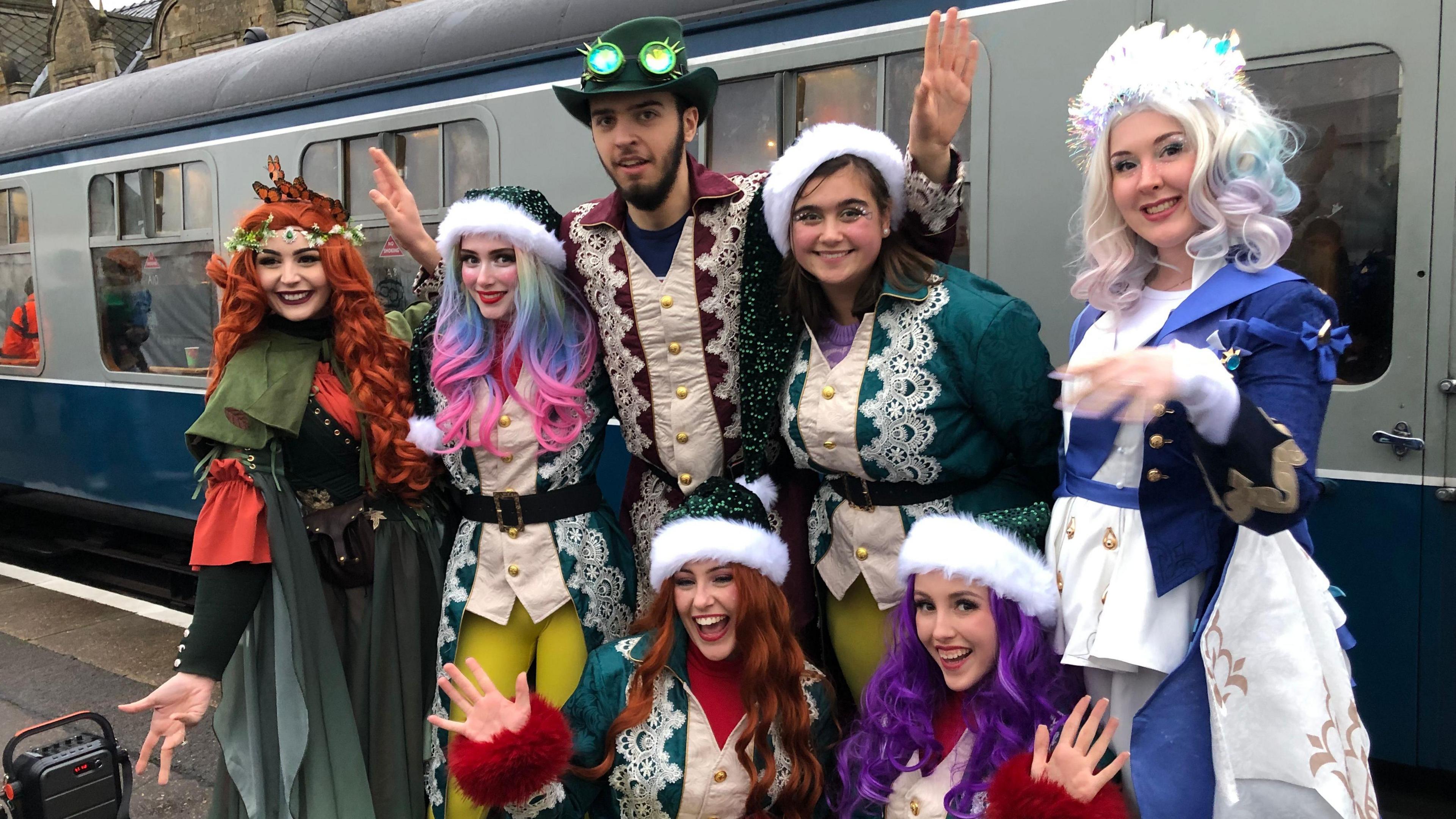 Seven cast members – six ladies and one man – dressed as elves and princesses. Behind them is one of the railway carriages