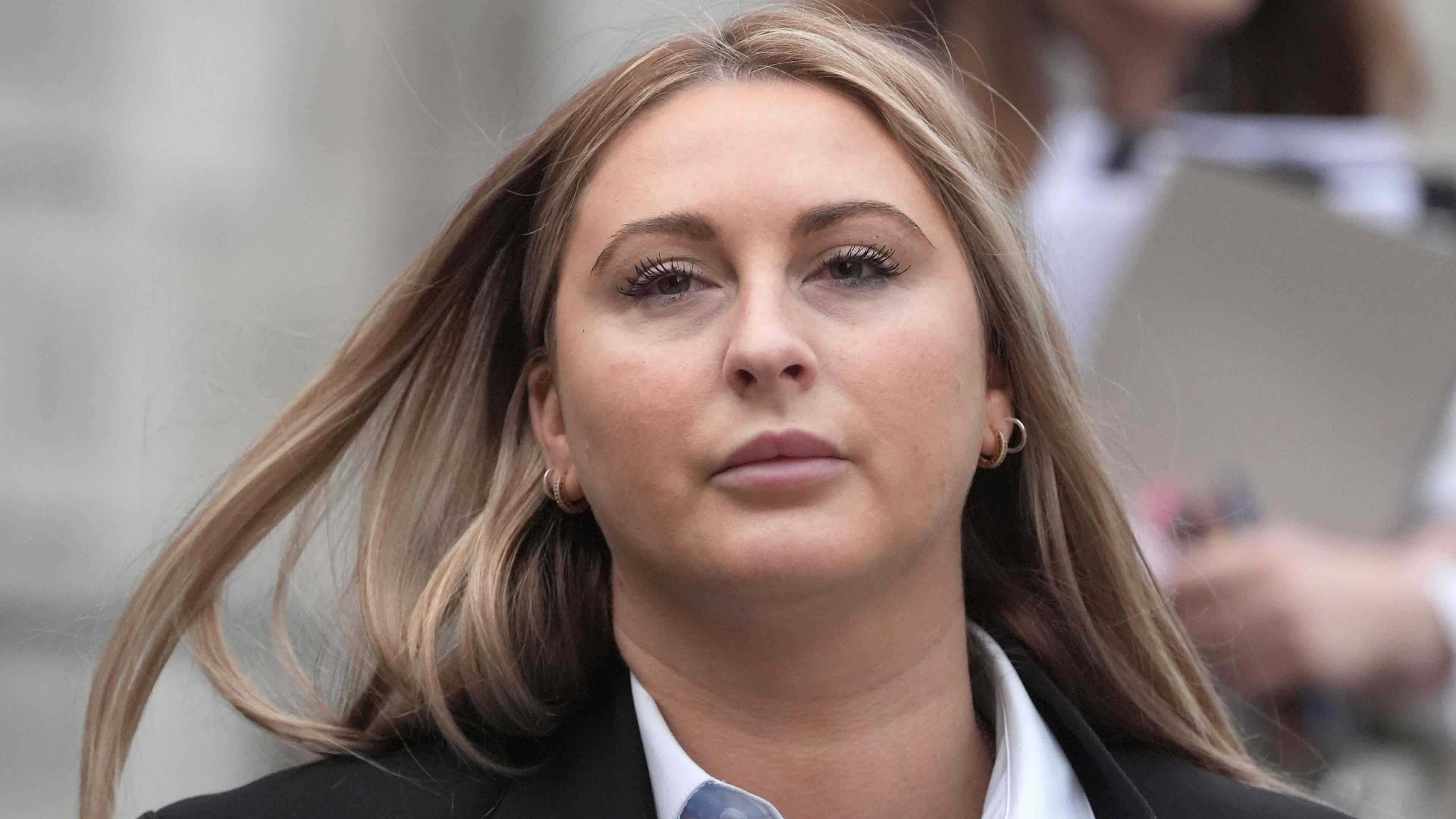 Nikita Hand is wearing a black jacket and a white shirt. She has fair hair and is wearing hoop earrings. She's outside a courthouse. There is a woman out of focus behind her.