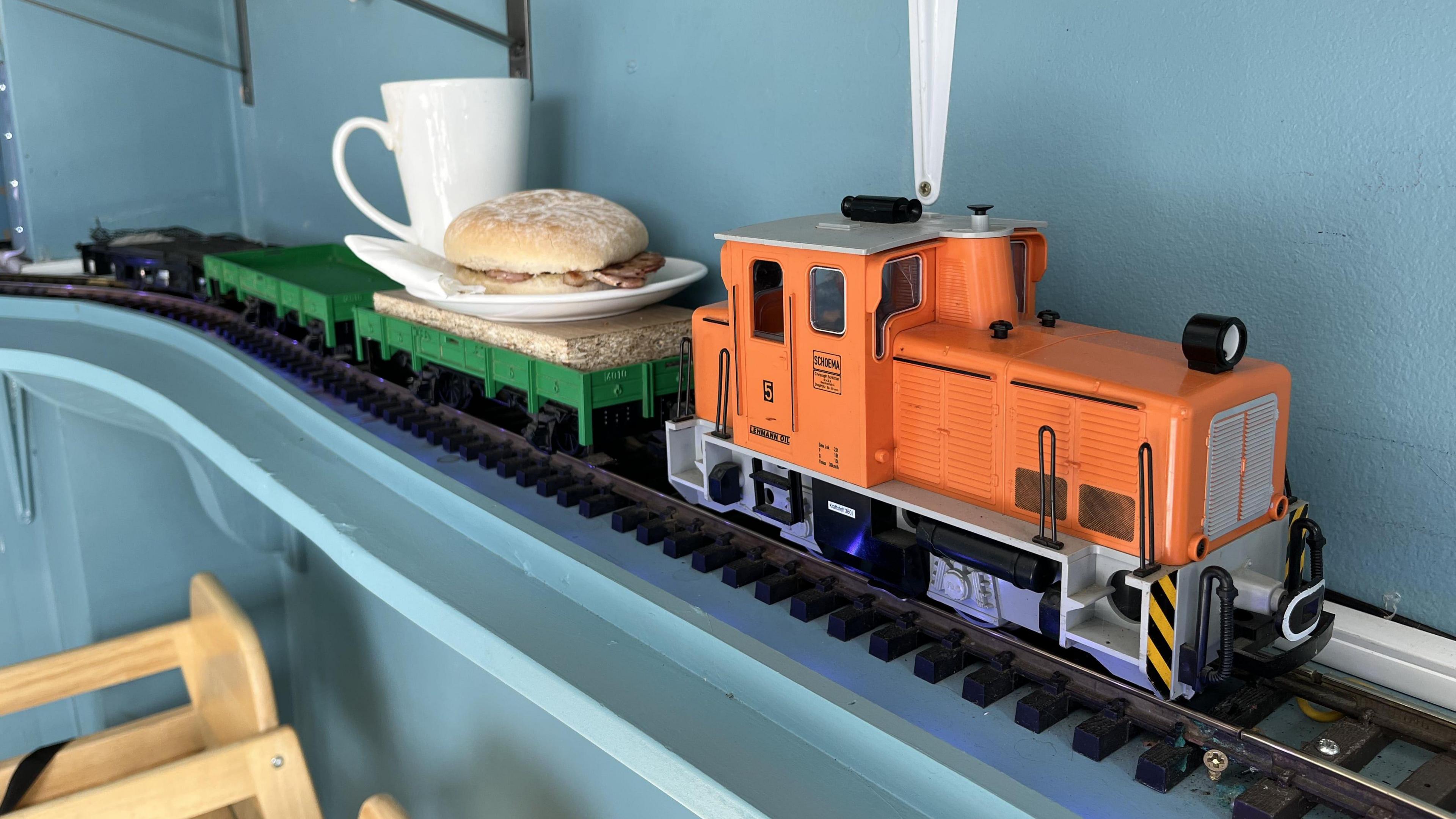 A small orange model train engine on tracks indoors pulling a bacon sandwich and a mug