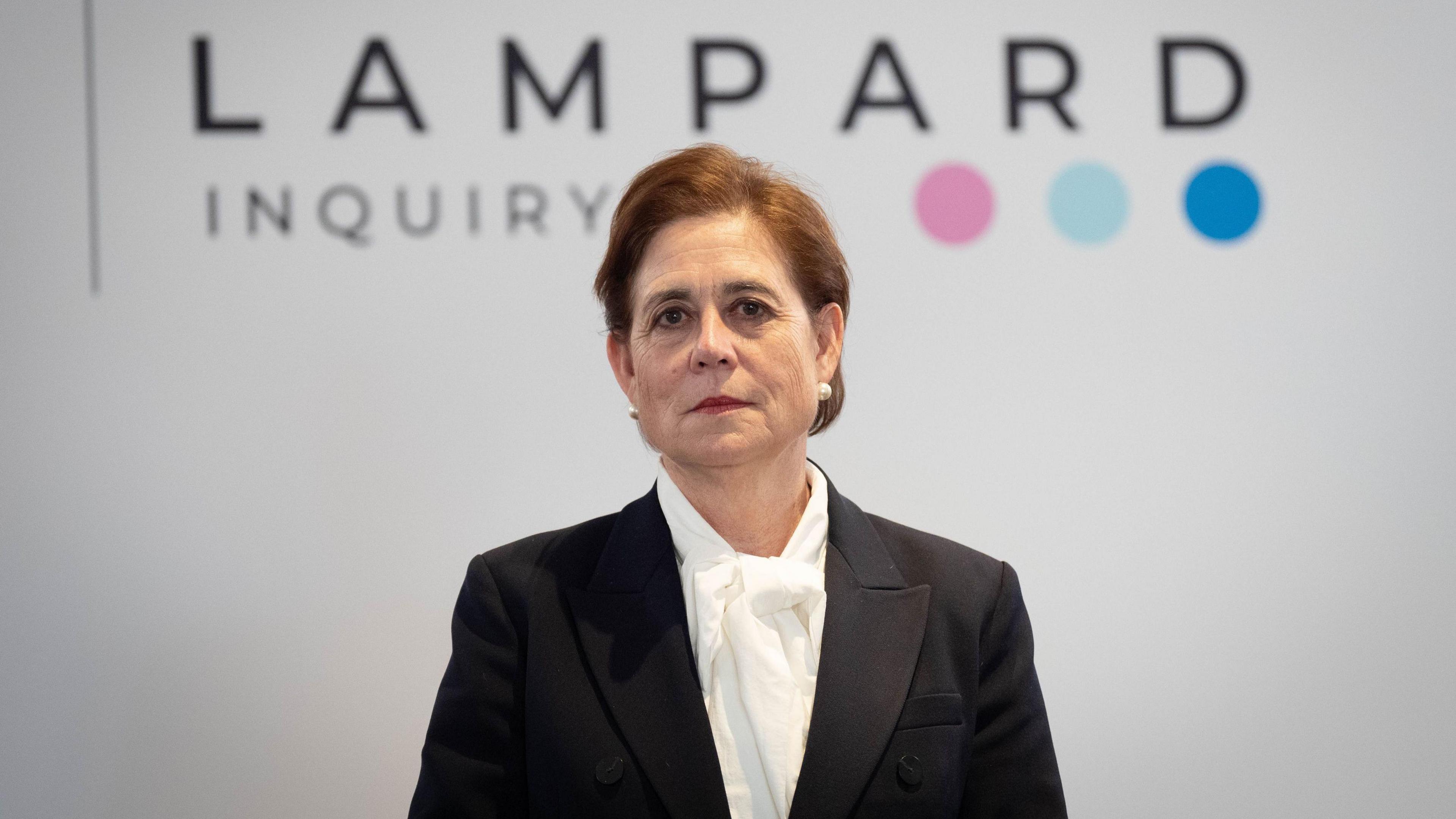 Baroness Lampard wears a black blazer jacket over a white blouse. She has dark brown short cropped hair and is looking straight-faced at the camera. She stands in front of a white backdrop which says the words "Lampard Inquiry" on it as well as three coloured dots