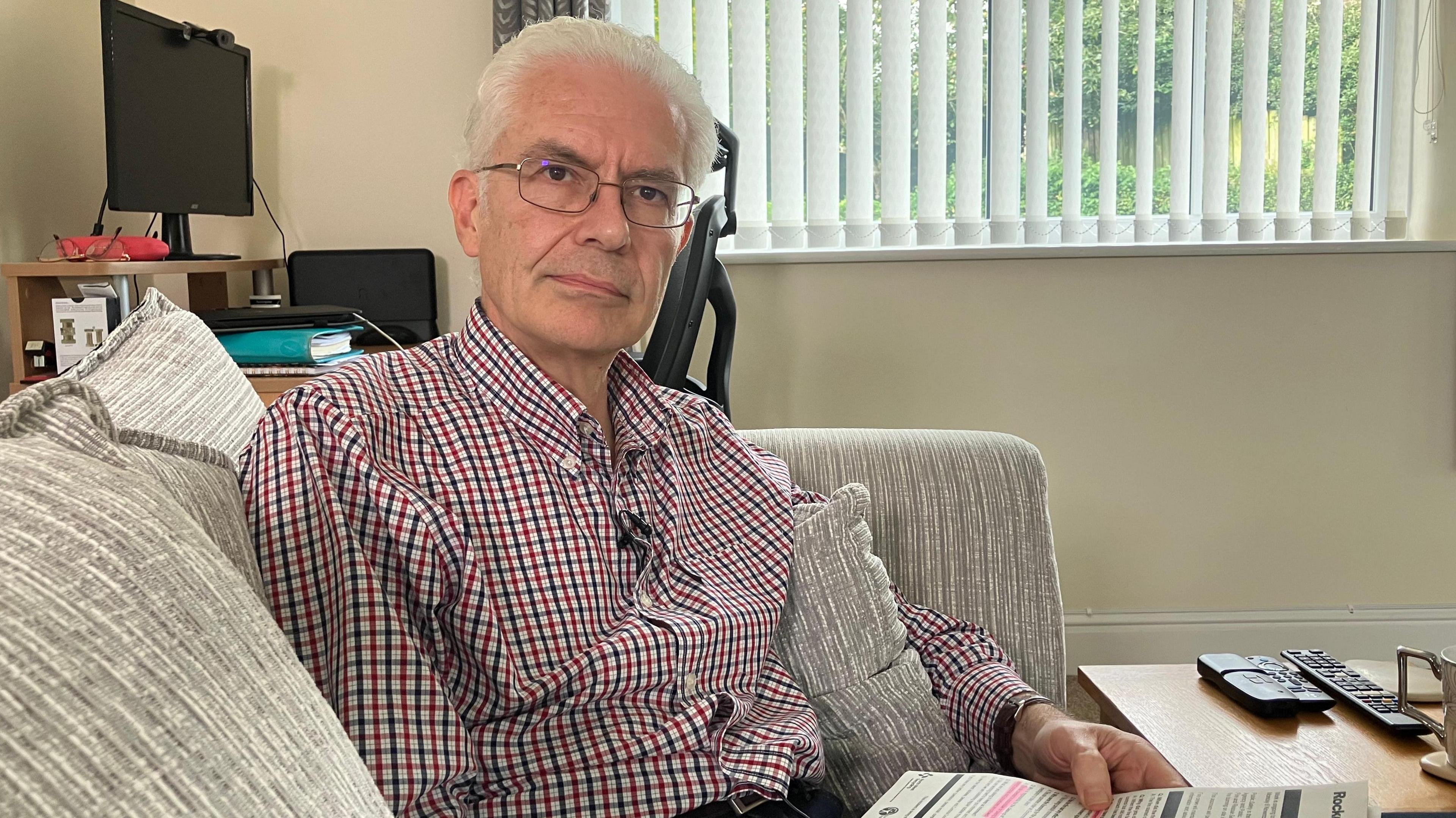 Campaigner David Mudge sitting on a sofa with a window behind him that has slatted horizontal blinds.