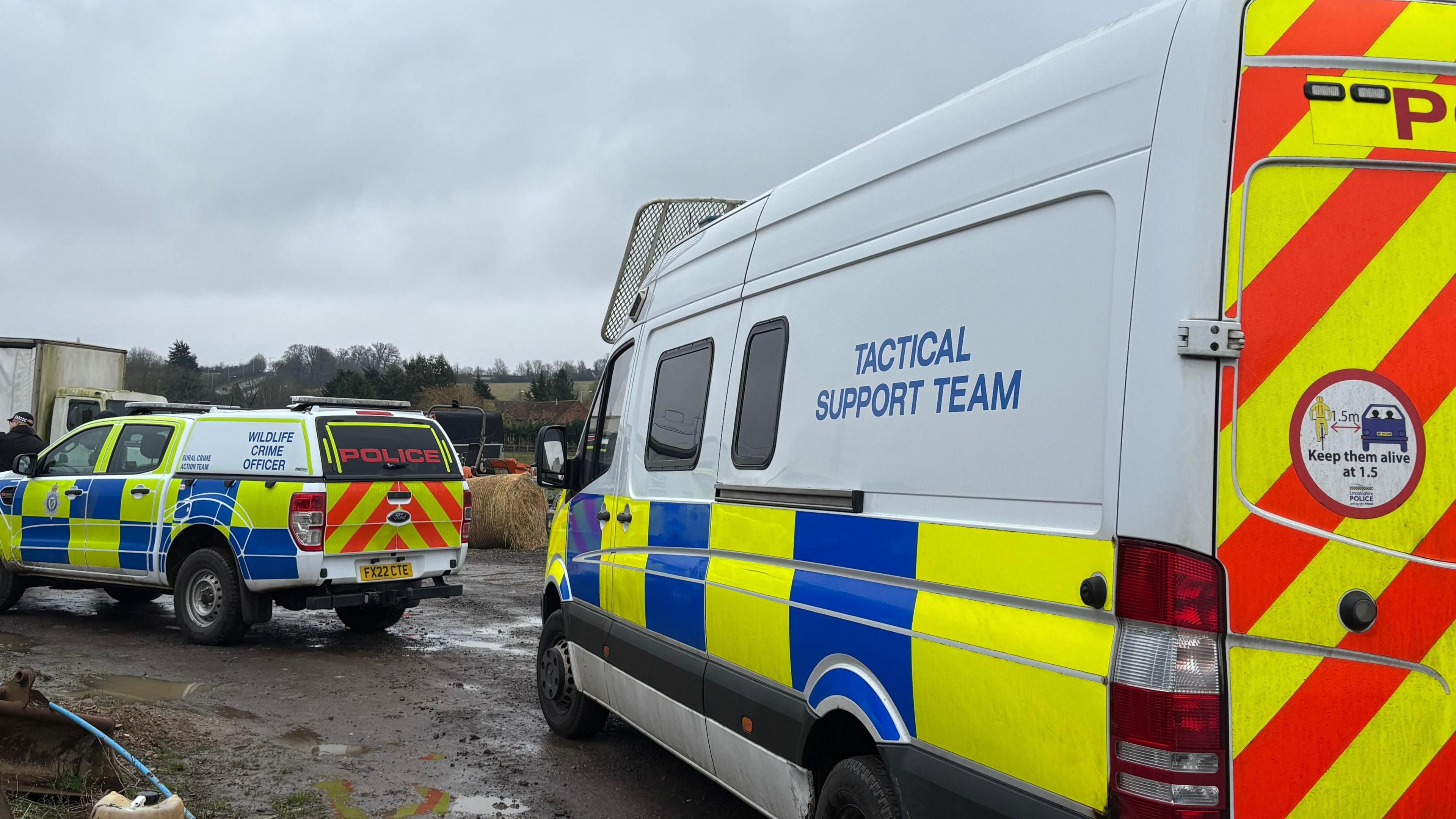 Two police tactical support vehicles are parked on what looks to be a farm. The ground is muddy with some puddles and there are fields and hay bales around them.