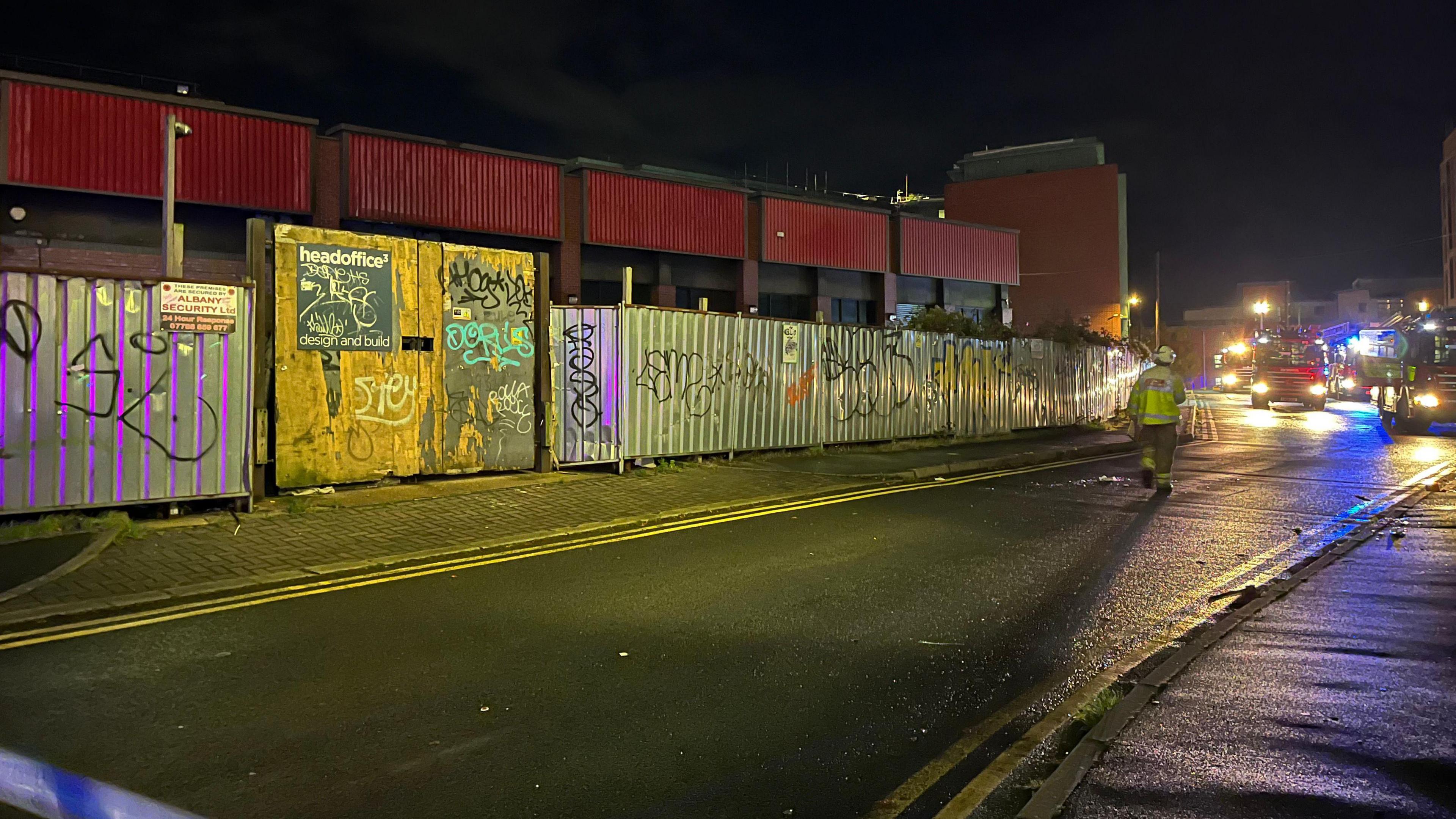 A police cordon around a disused commercial building in Nottingham. The building, once a Staples office supplies shop, is boarded up with metal fencing. A number of fire service vehicles can be scene in the background while a firefighter crosses the road. It is night and dark outside. Blue lights can be scene from emergency service vehicles.