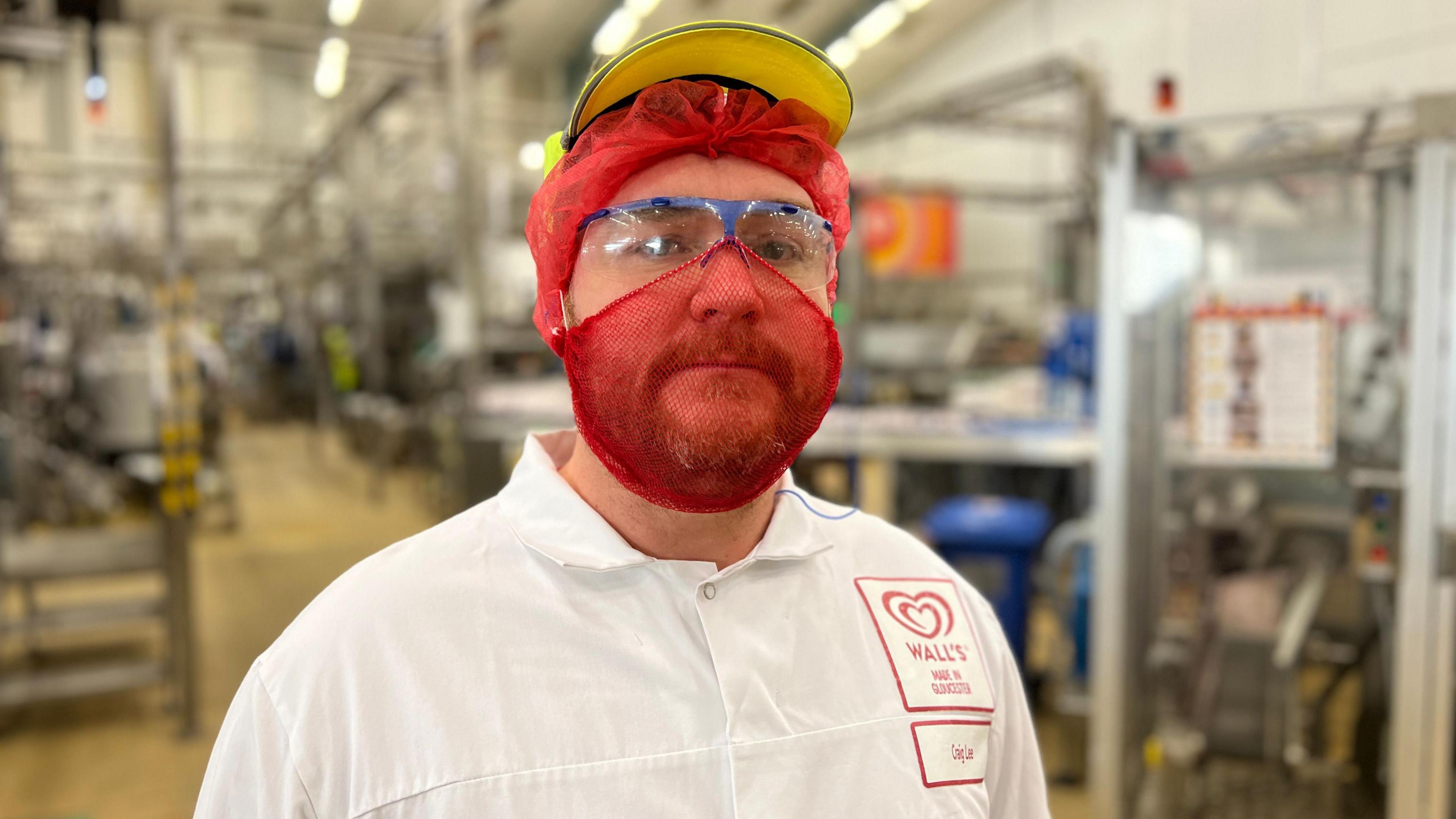 Callum is standing in the factory wearing protective equipment. He is wearing a red beard- and hair net and a yellow cap. 