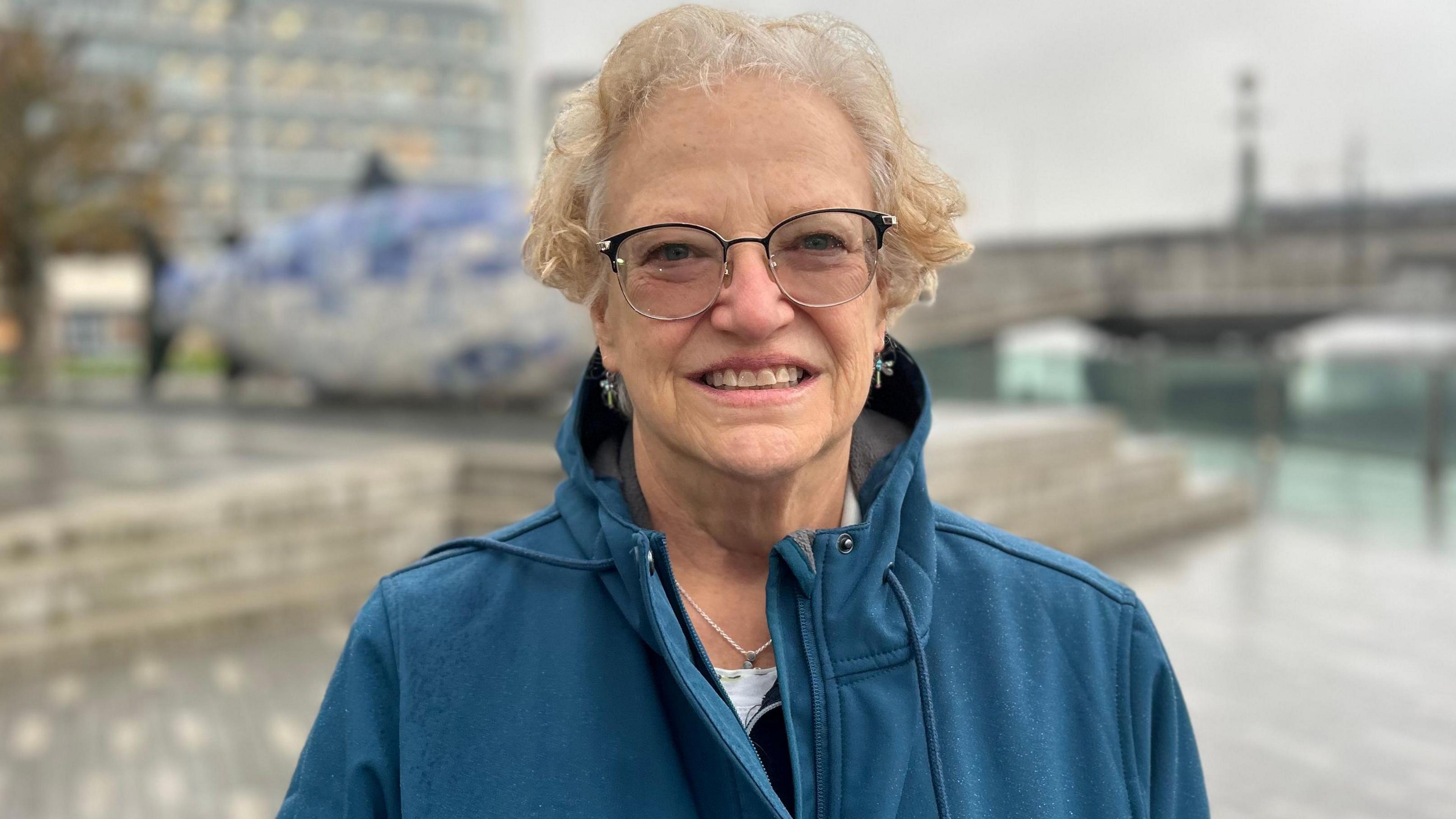 Lesly wears glasses and has insect earrings on. She's smiling. Belfast's Big Fish sculptor is behind her. The streets are wet with rain, so she's wearing a green rain coat. She has short fair hair.