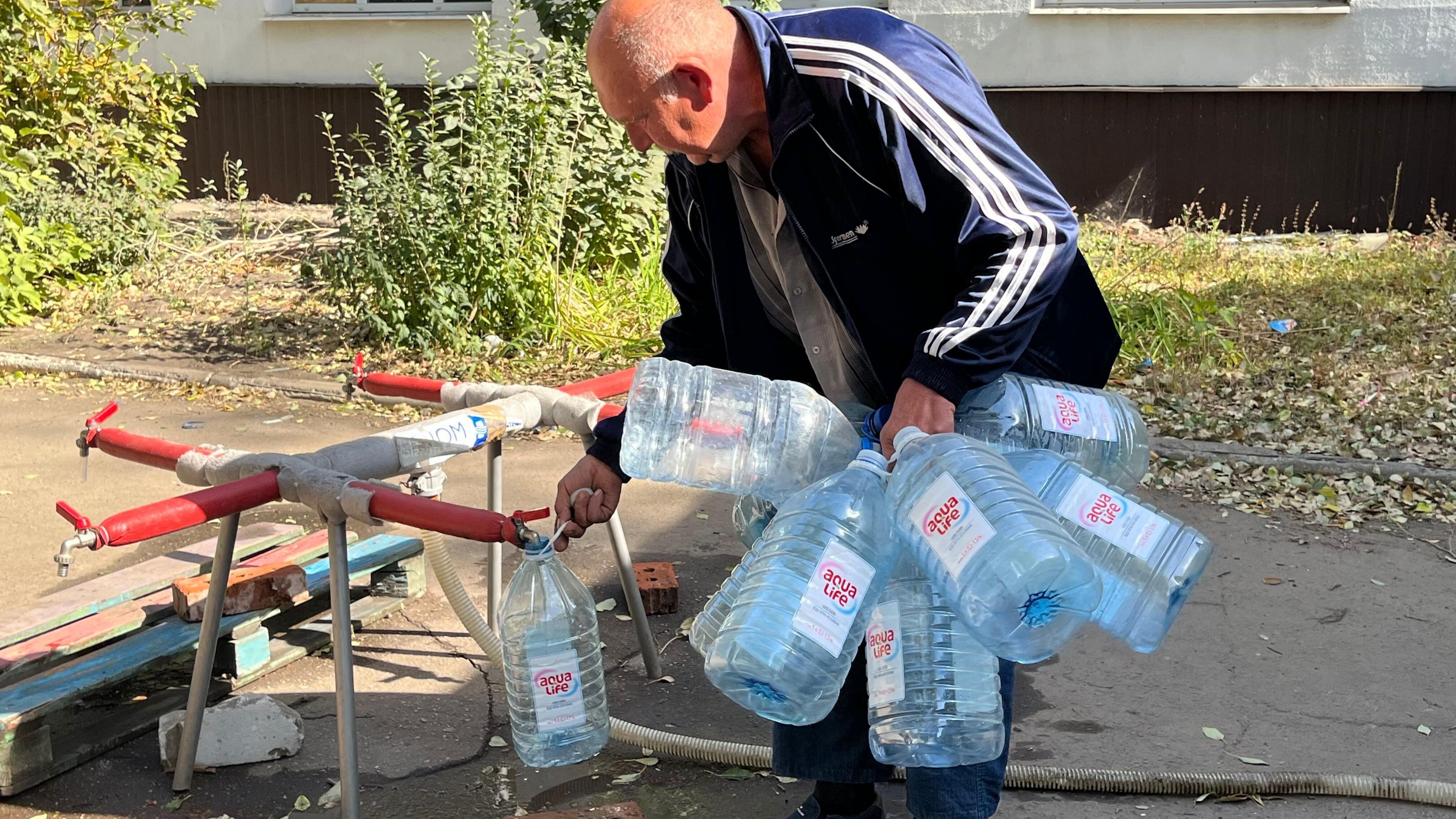 A middle-aged man in a blue jacket fill ups water canisters from a makeshift arrangements of taps outdoors on a sunny day