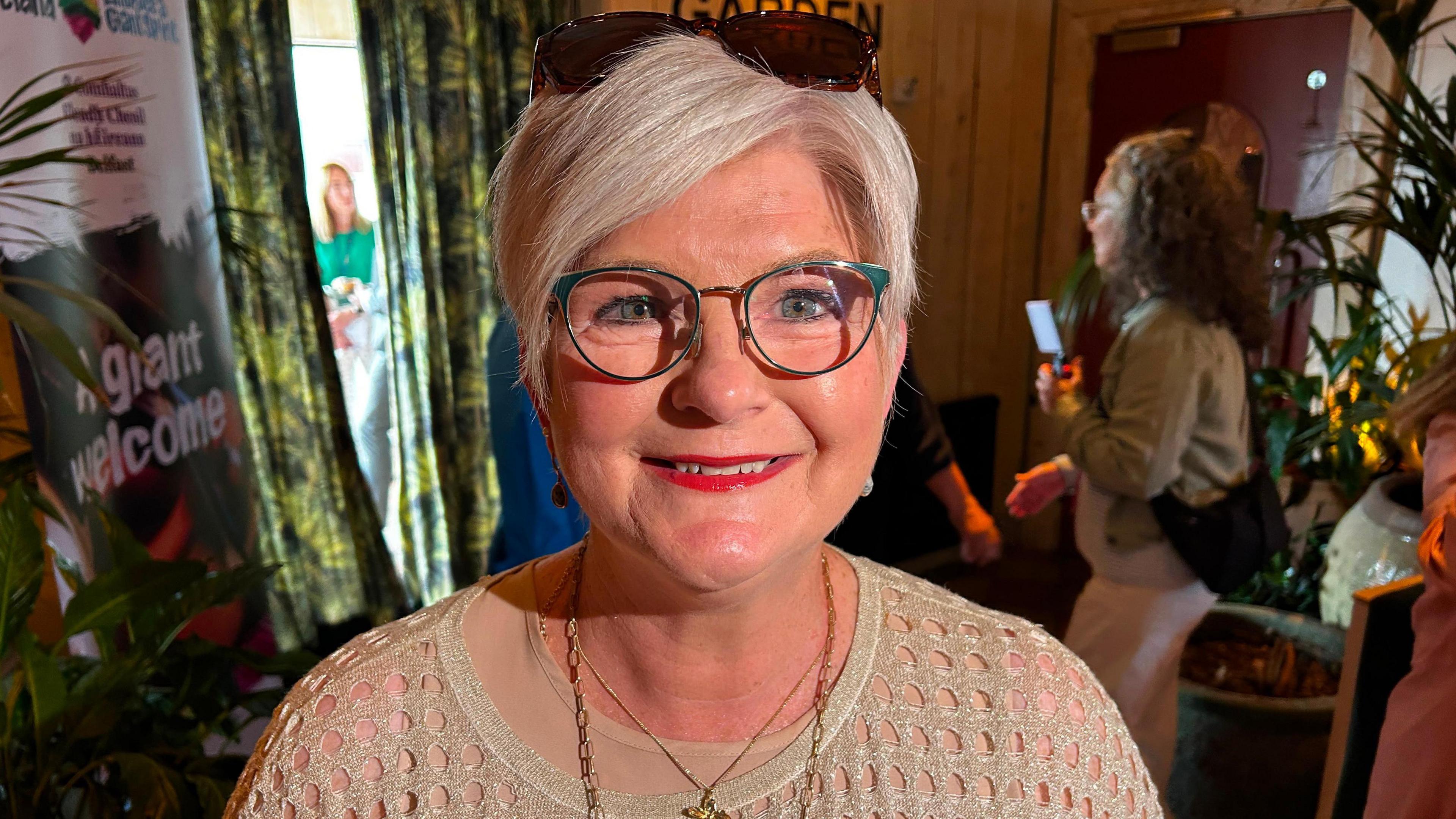 A woman in a light brown jumper wearing necklaces. She has white short hair and wears glasses and red lipstick. There are sunglasses on her head.