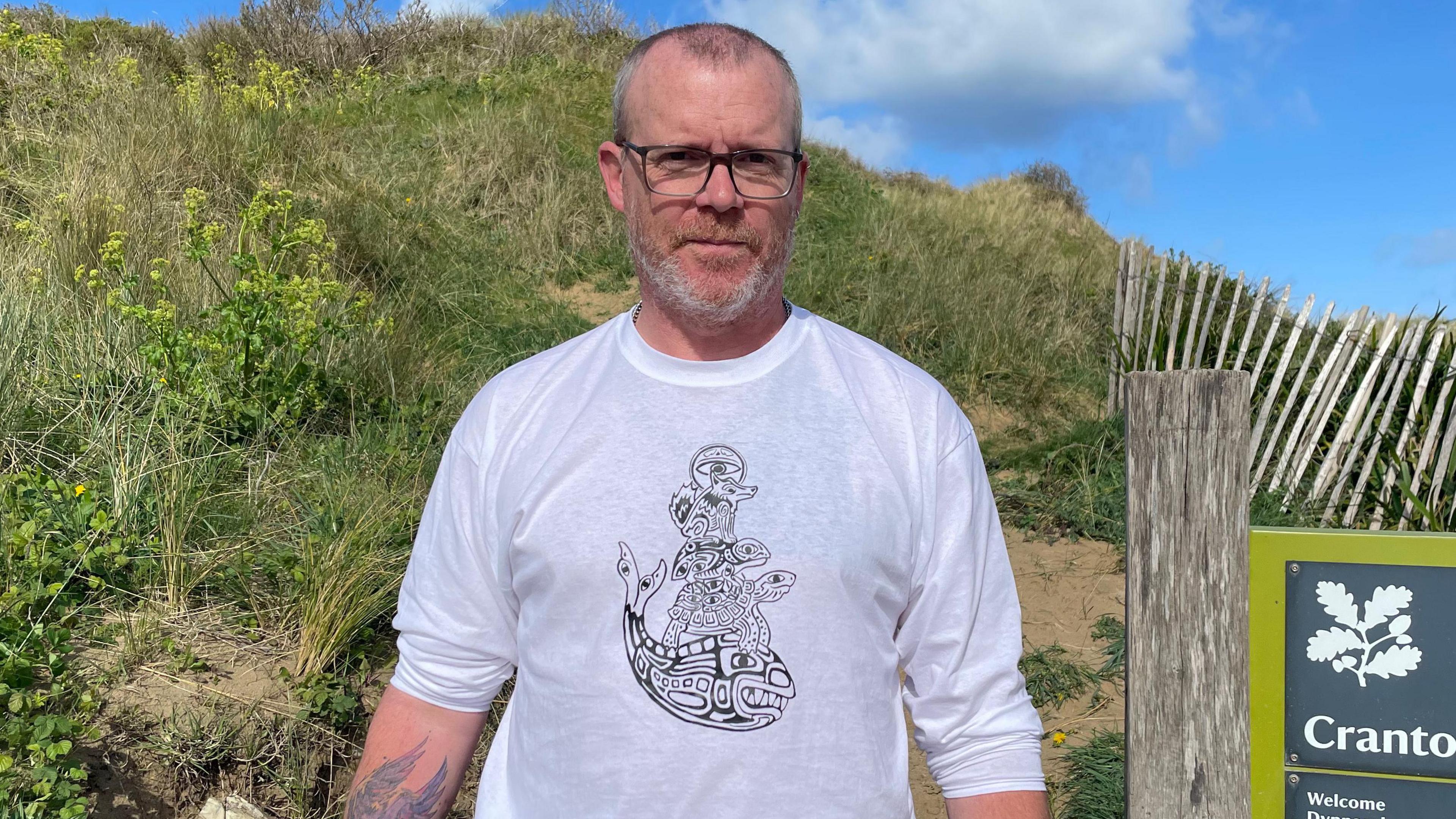 Matt Cole is smiling as he stands in front of a sand dune on a sunny day. He is wearing glasses, a white long-sleeved T-shirt with a black design on it and he has a tattoo of wings on his right forearm. Next to him part of a sign can be seen which has a National Trust logo and the letters Cranto and Welcome.