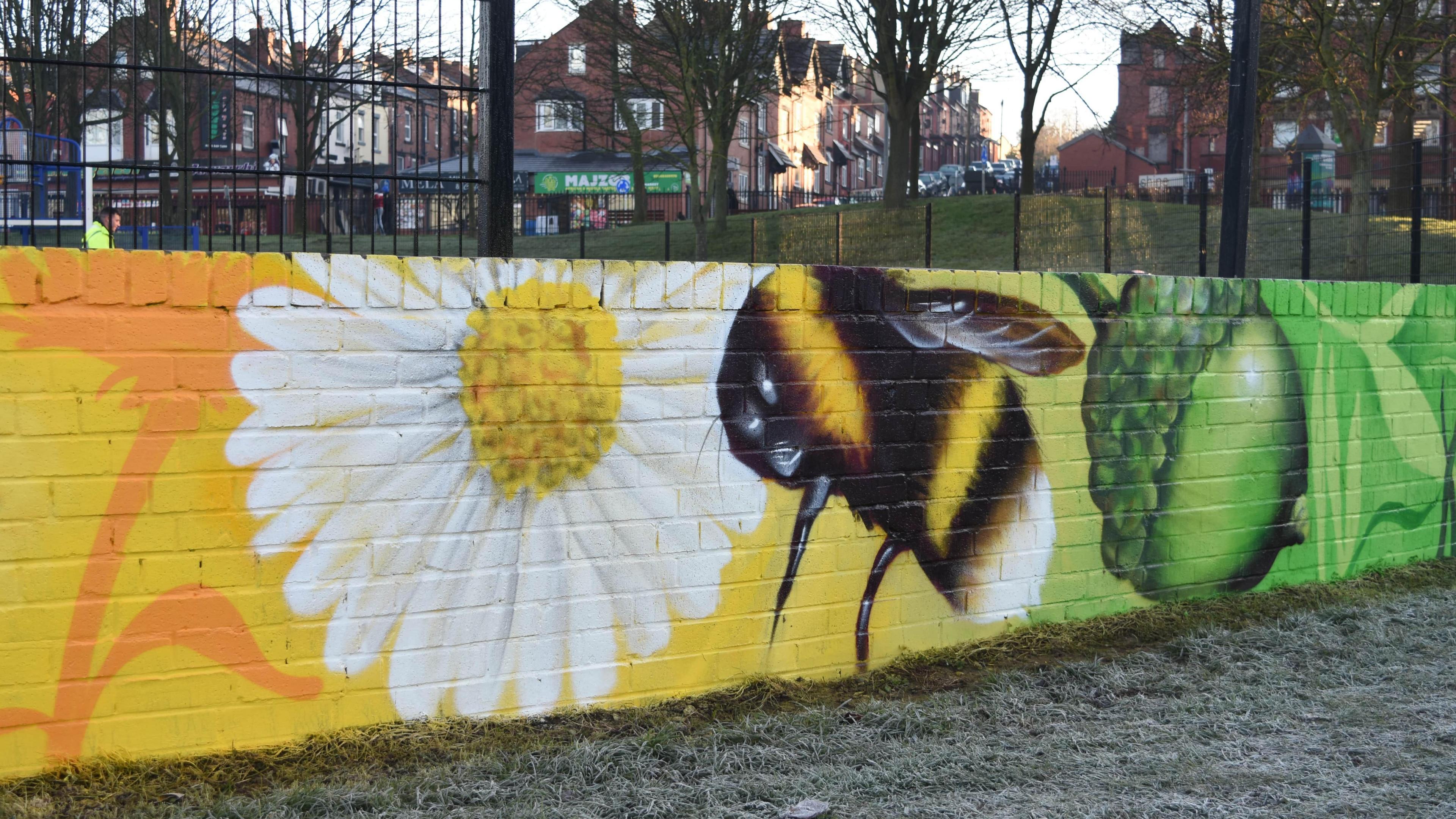 A brightly-coloured mural on a wall of about five foot in a park. It features a bee and a daisy against a yellow backdrop and an acorn against a green backdrop.