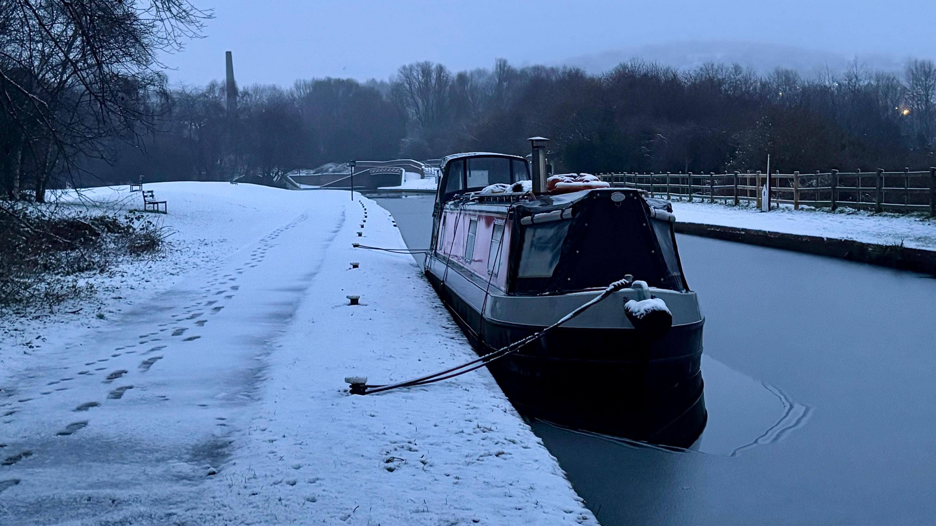 A barge on a frozen canal in Netherton, West Midlands. The tow path has a dusting of snow with two pairs of footprints. Woodland surrounds the canal and the sky is grey. 