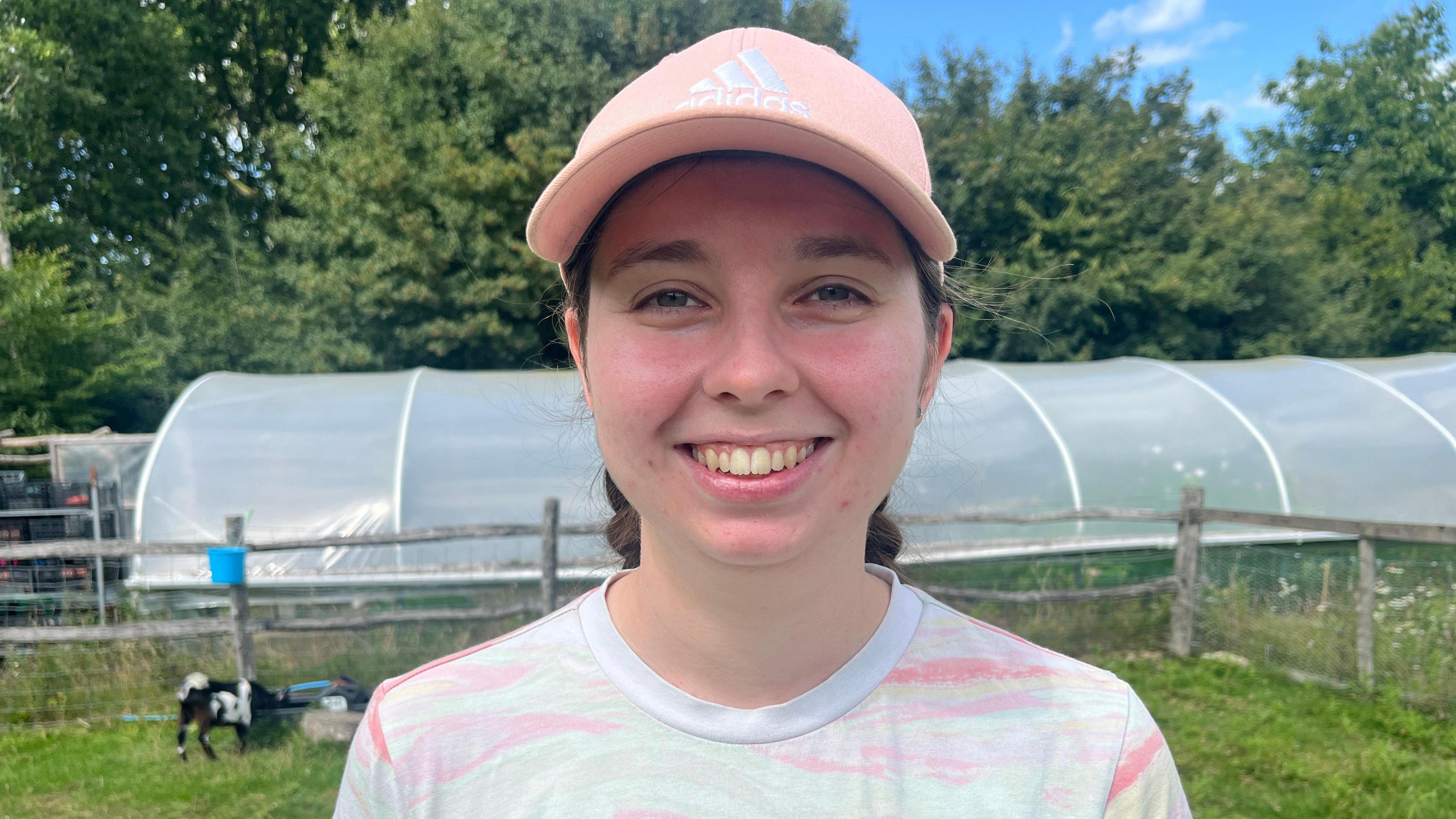 Chloe is standing in a field wearing a pink cap and smiling to camera.