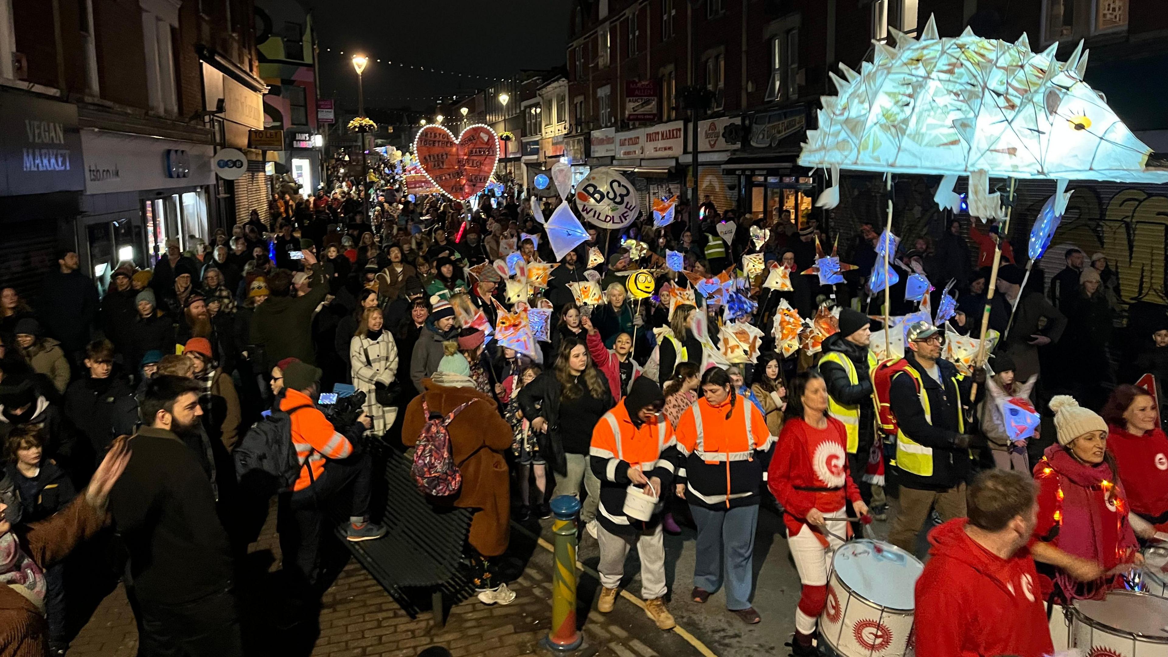 Participants in the Bedminster Winter Lantern Parade walk down East Street. The photograph is taken at night and hundreds of people are visible, many of them carrying colourful, illuminated lanterns.