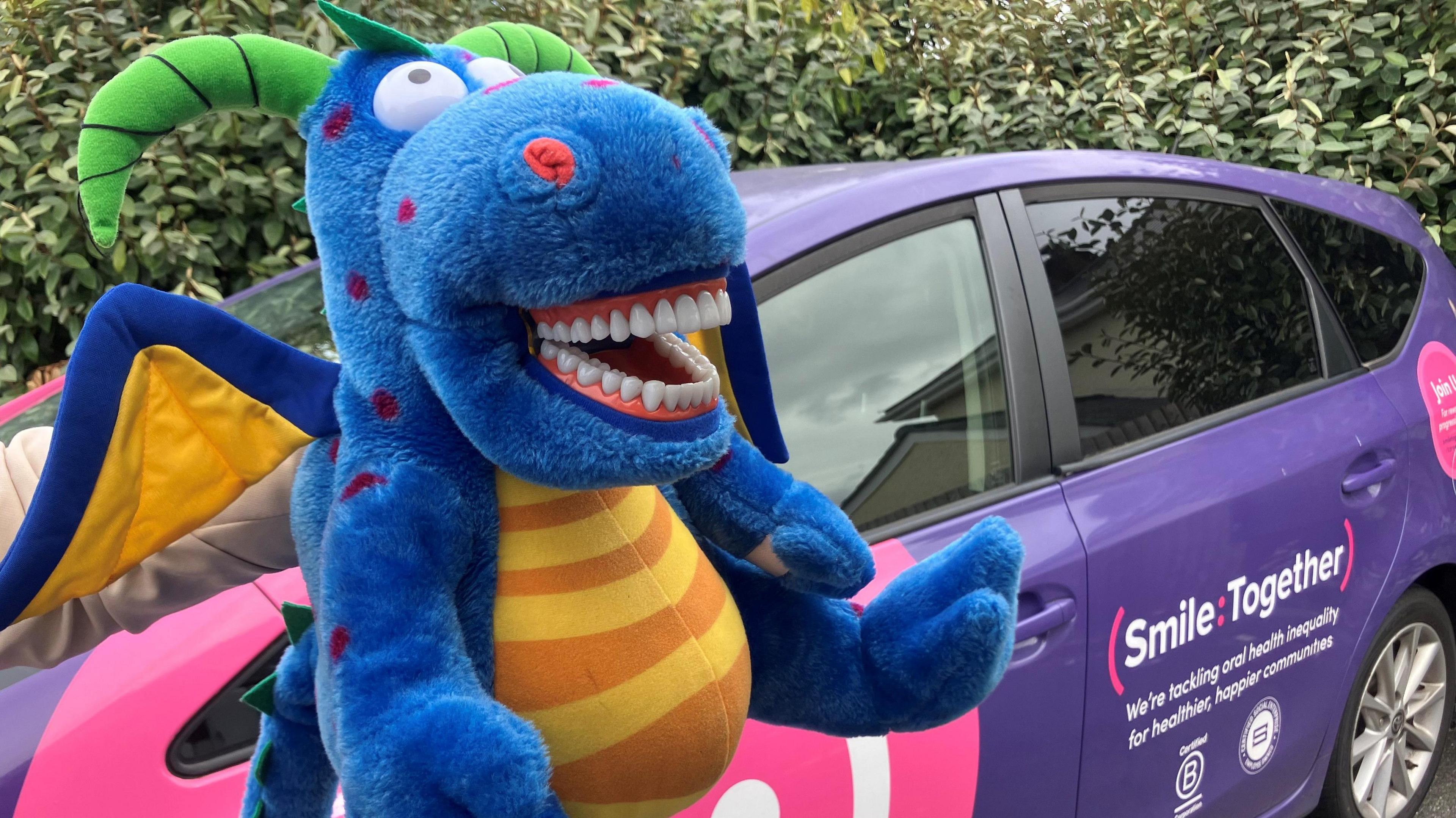 A toy dragon is held up. He has a big set of human-looking teeth which are white and straight.