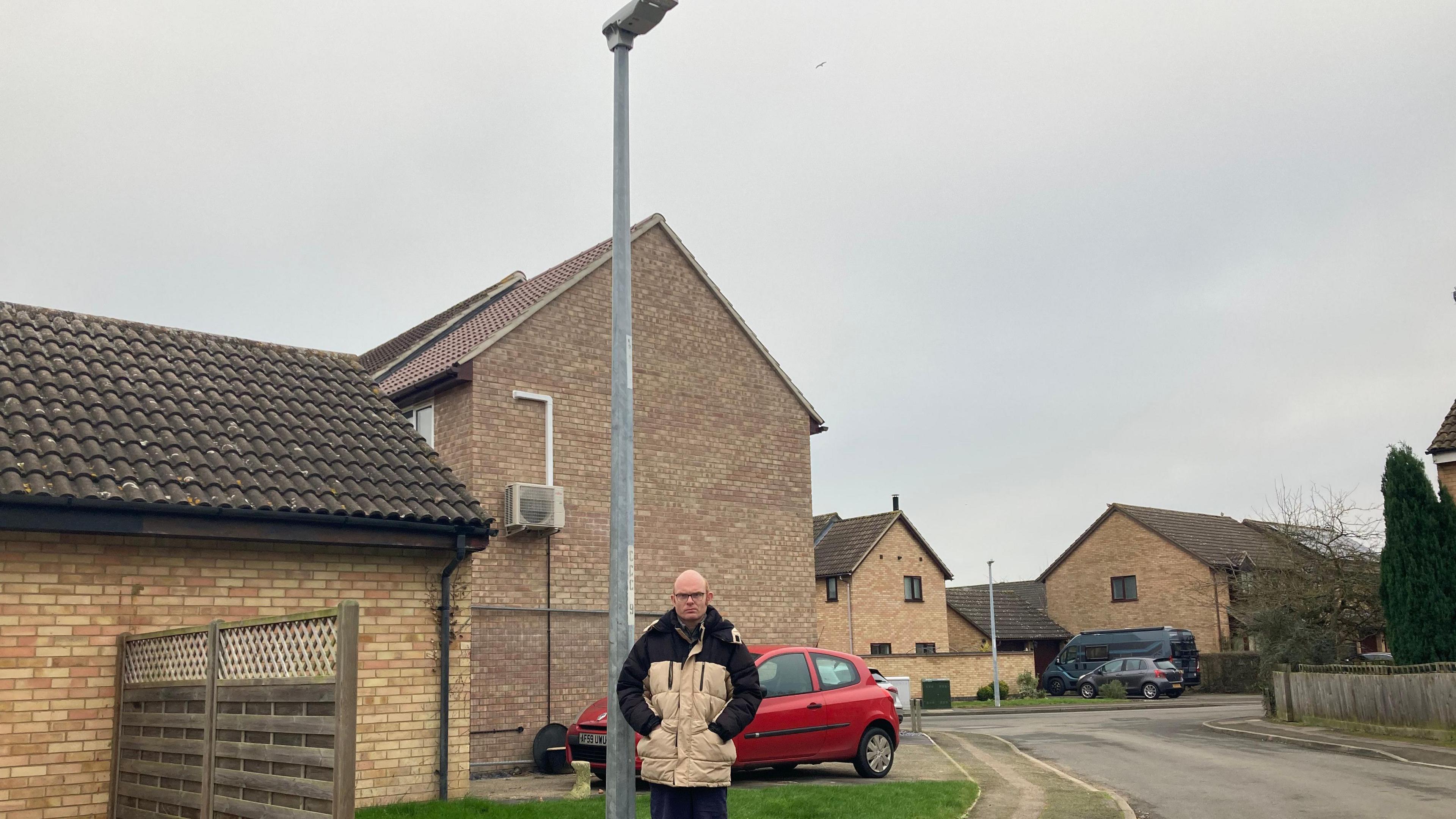 James Windle, a bald man wearing a black and beige coat, stands underneath a lamp post in a street.