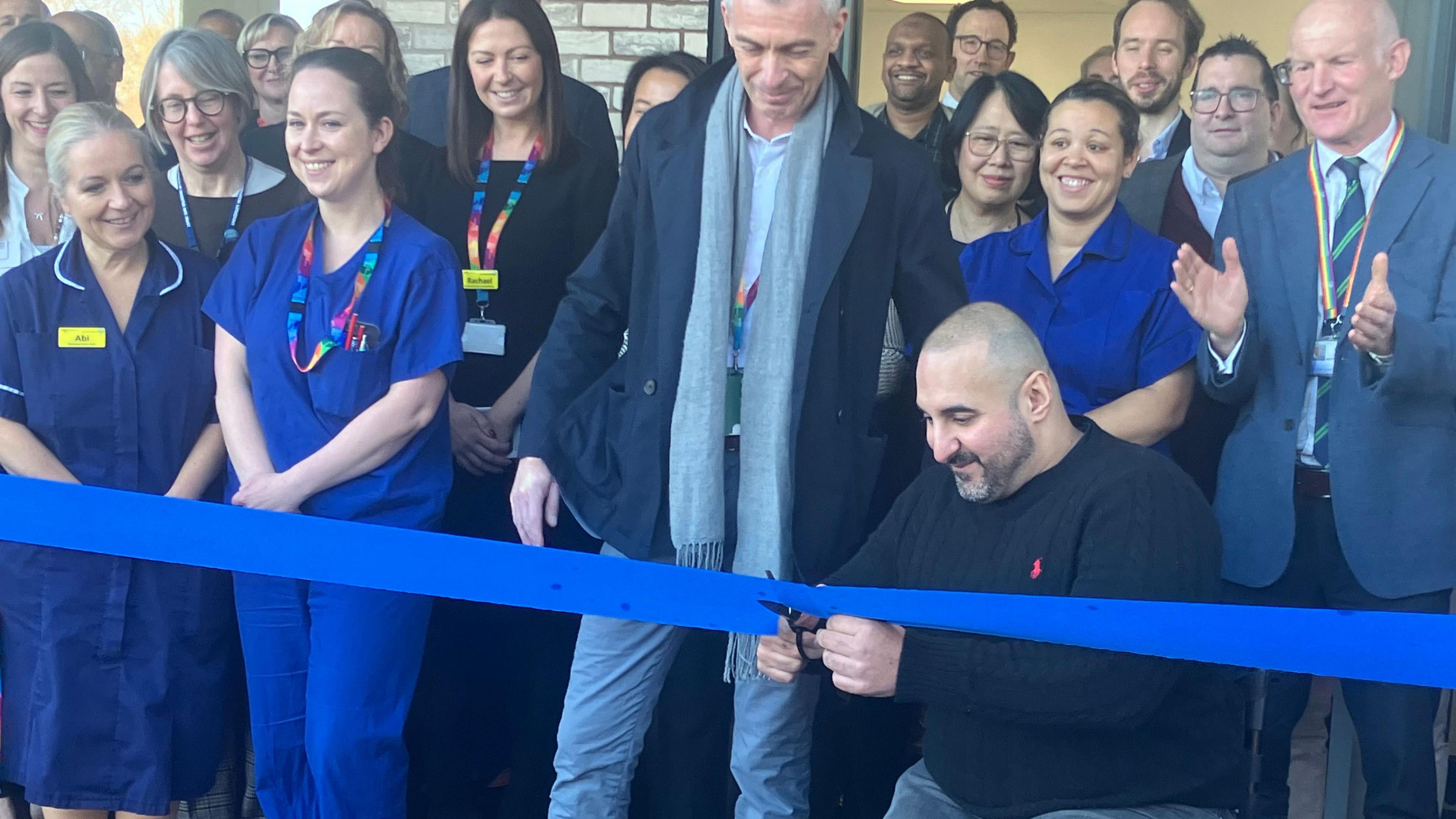 British powerlifter, Ali Jawad, cutting a blue ribbon, officially opening the endoscopy centre at Leicester General Hospital on Tuesday. The are people standing with him smiling and clapping.