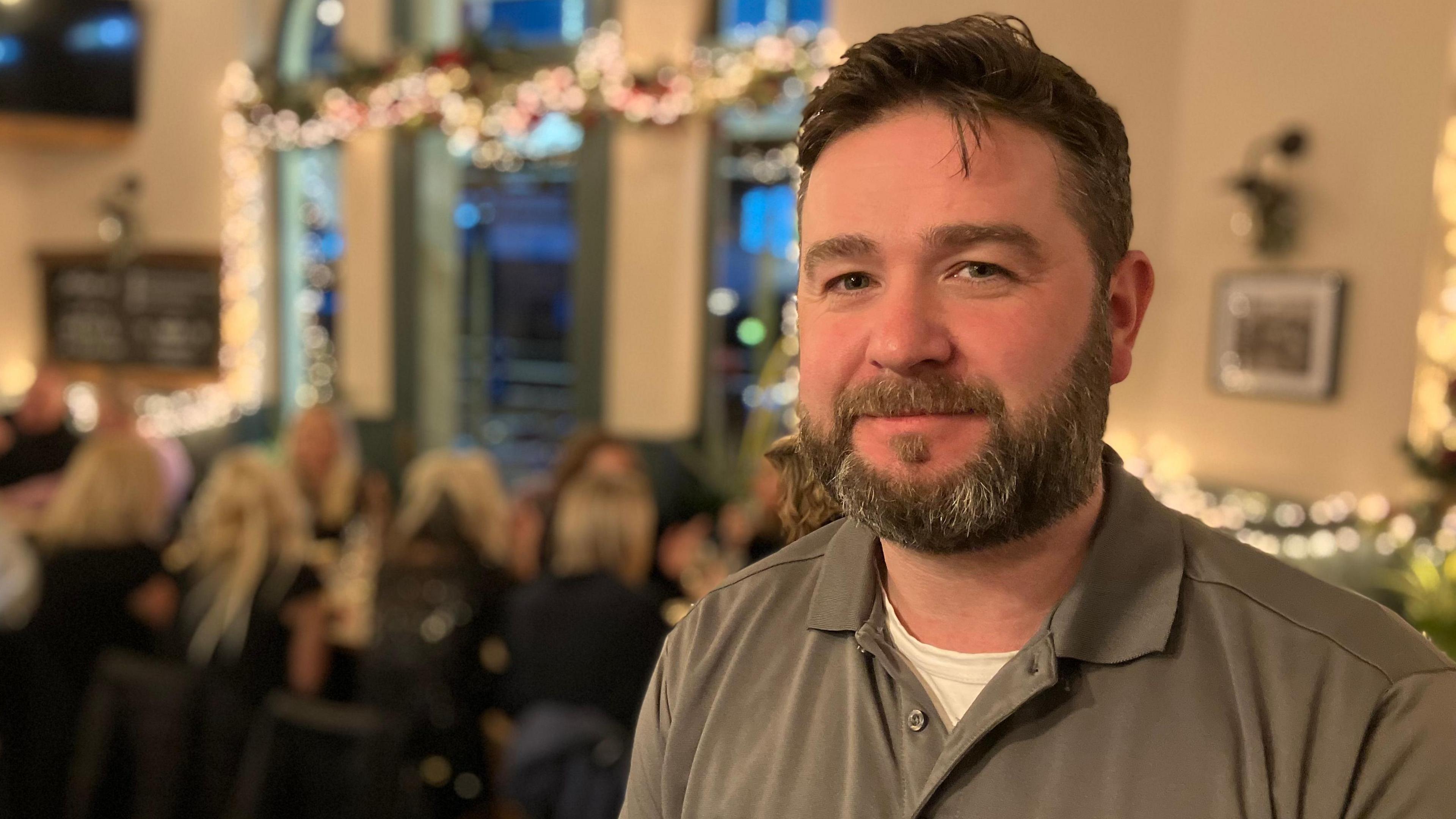 A man stands in a busy restaurant. There is dining table in the background that has around a dozen people sitting around it.  There are Christmas decorations and festive lights around the room.  