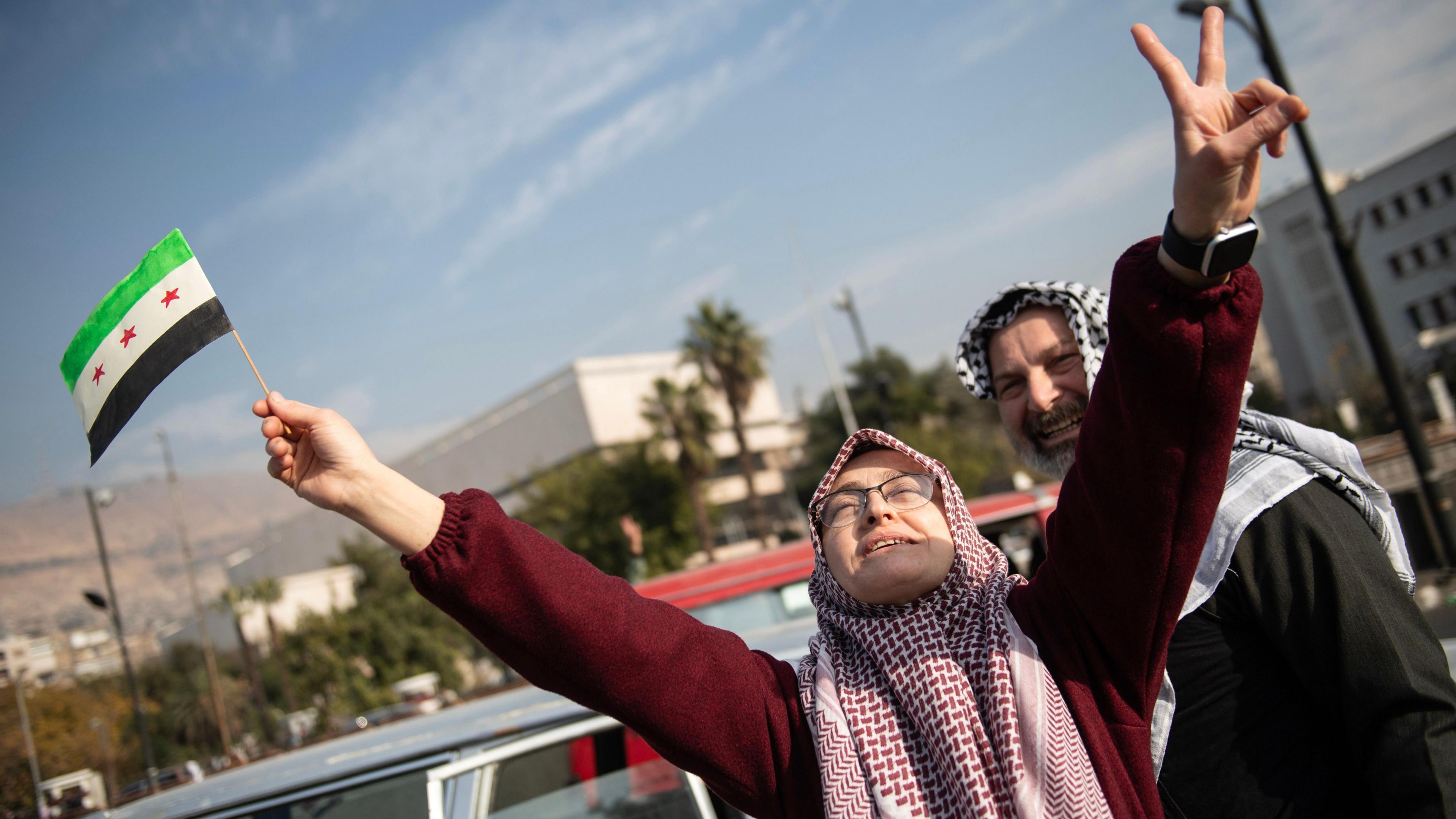 Woman in hijab celebrates the toppling of the Assad regime, face tilted up and her hands held up in the air. In one hand she holds the Syrian rebel flag. A man stands behind her smiling.