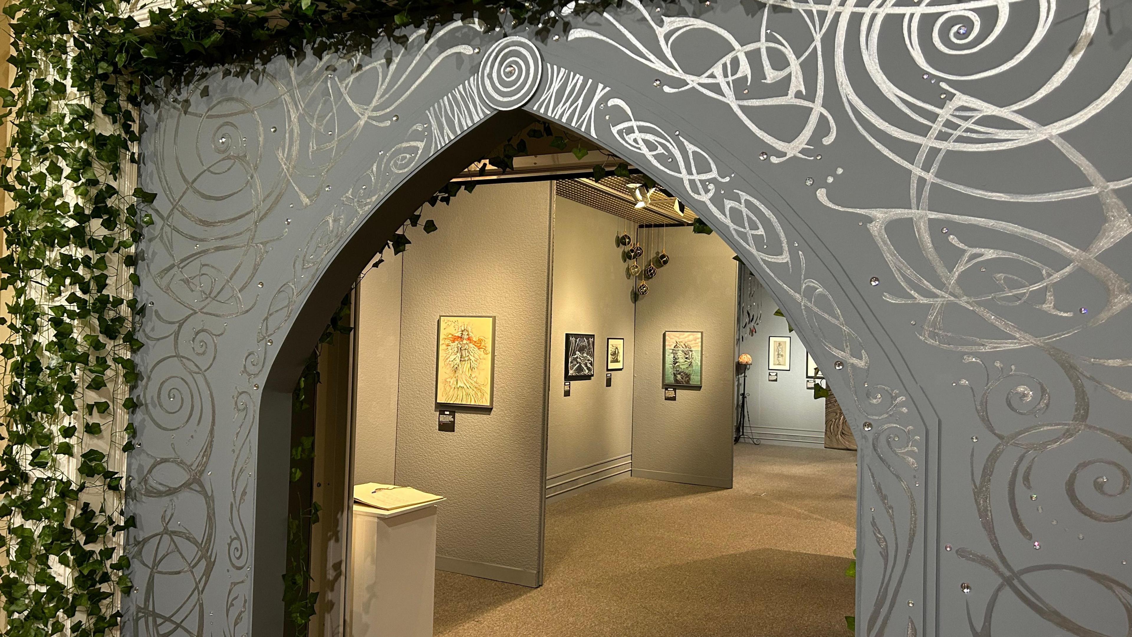 A mystical looking entrance with celtic designs painted on it and ivy around it, you can see the exhibition in the backgroun.