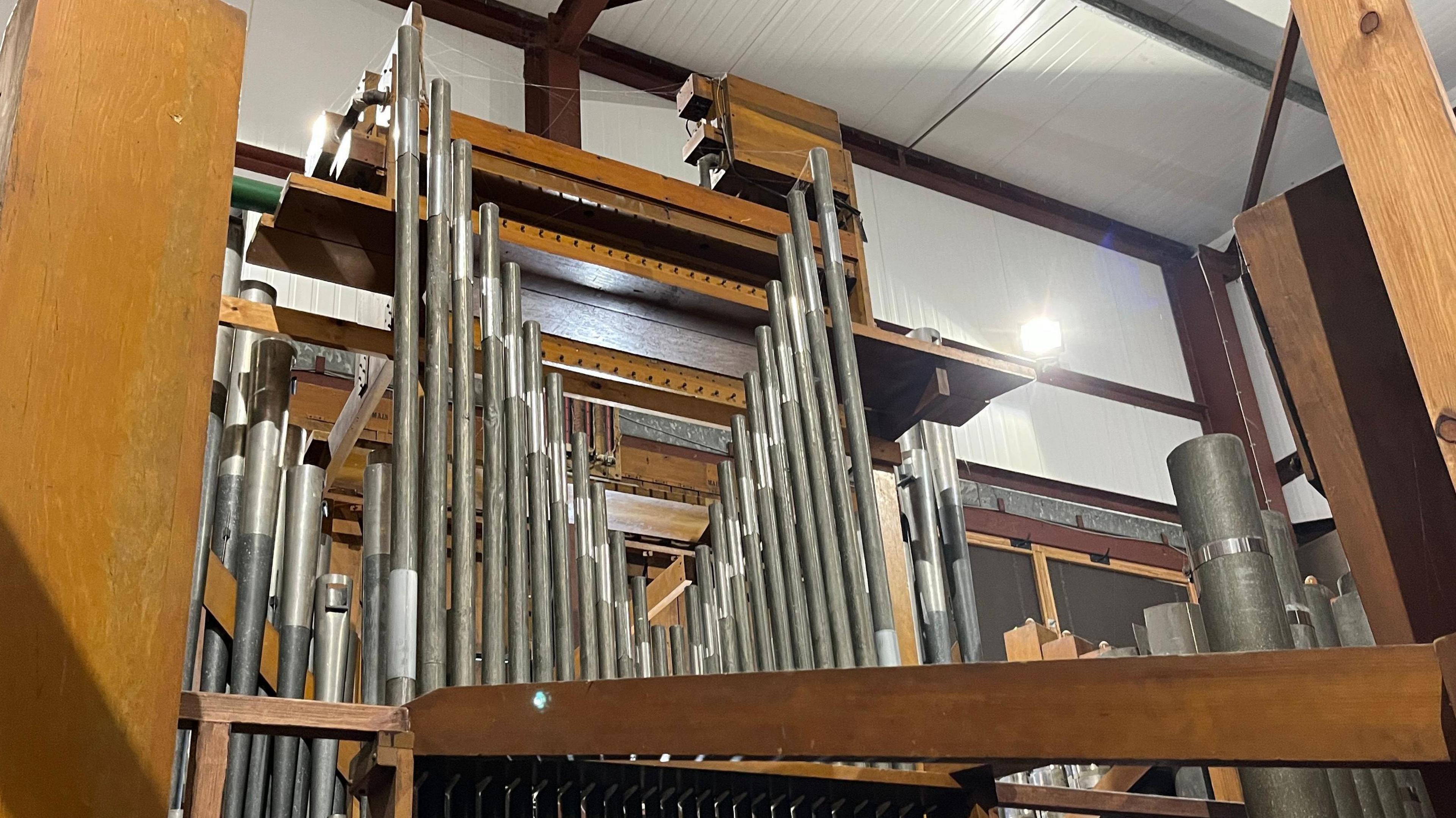 Close view of a Wurlitzer organ's pipes which are silver. Pieces of light brown wood are positioned around the organ, which go around the pipes. 