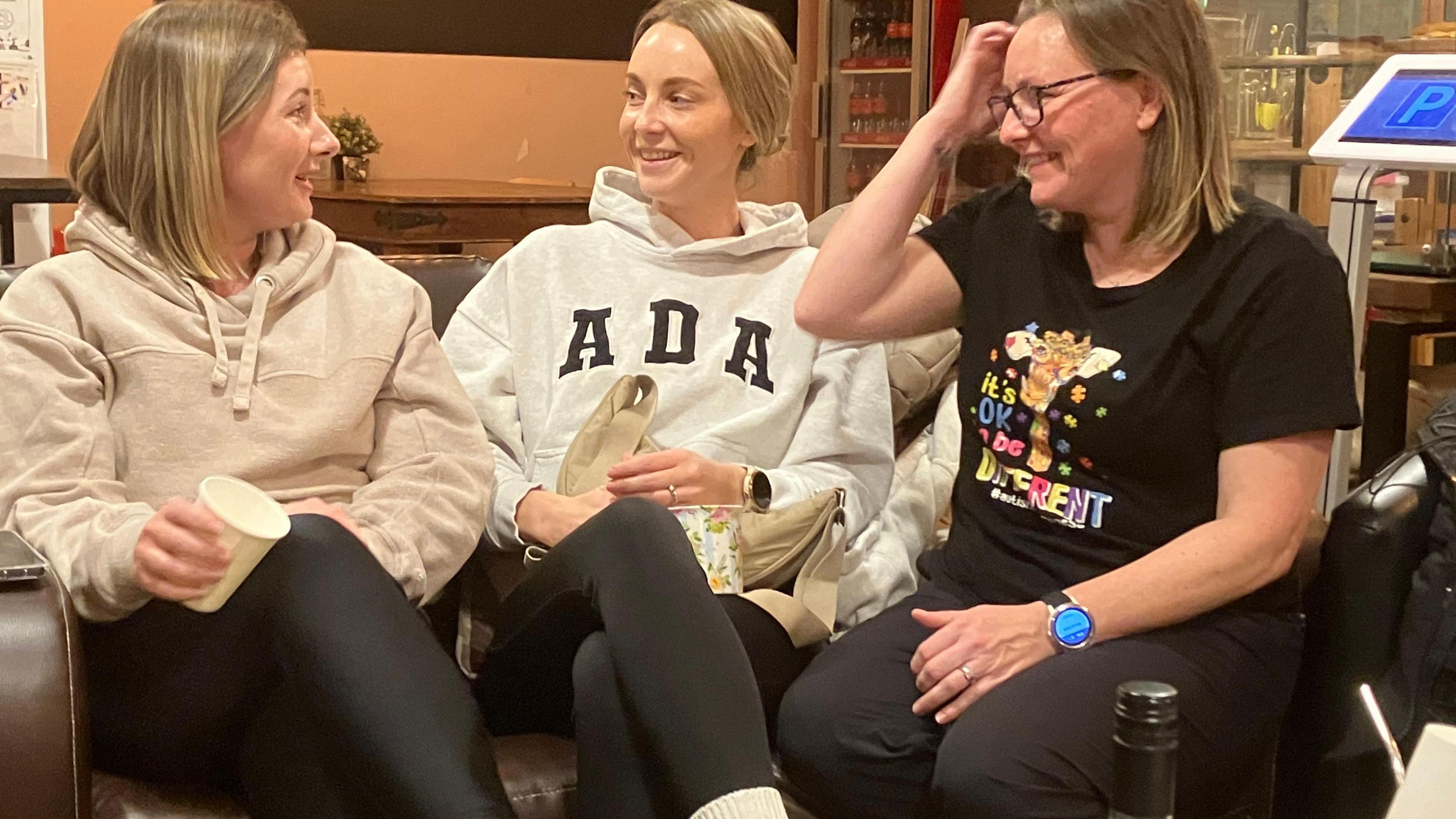 A picture of three women sat on a sofa. The one on the left has dark blonde shoulder length hair, a cream fleece and is holding a coffee cup in her right hand. In the middle is a blonde woman with her hair tied back, who is wearing a white fleece with the letters ADA on. On the right is a woman with light brown shoulder length hair with a black t-shirt.