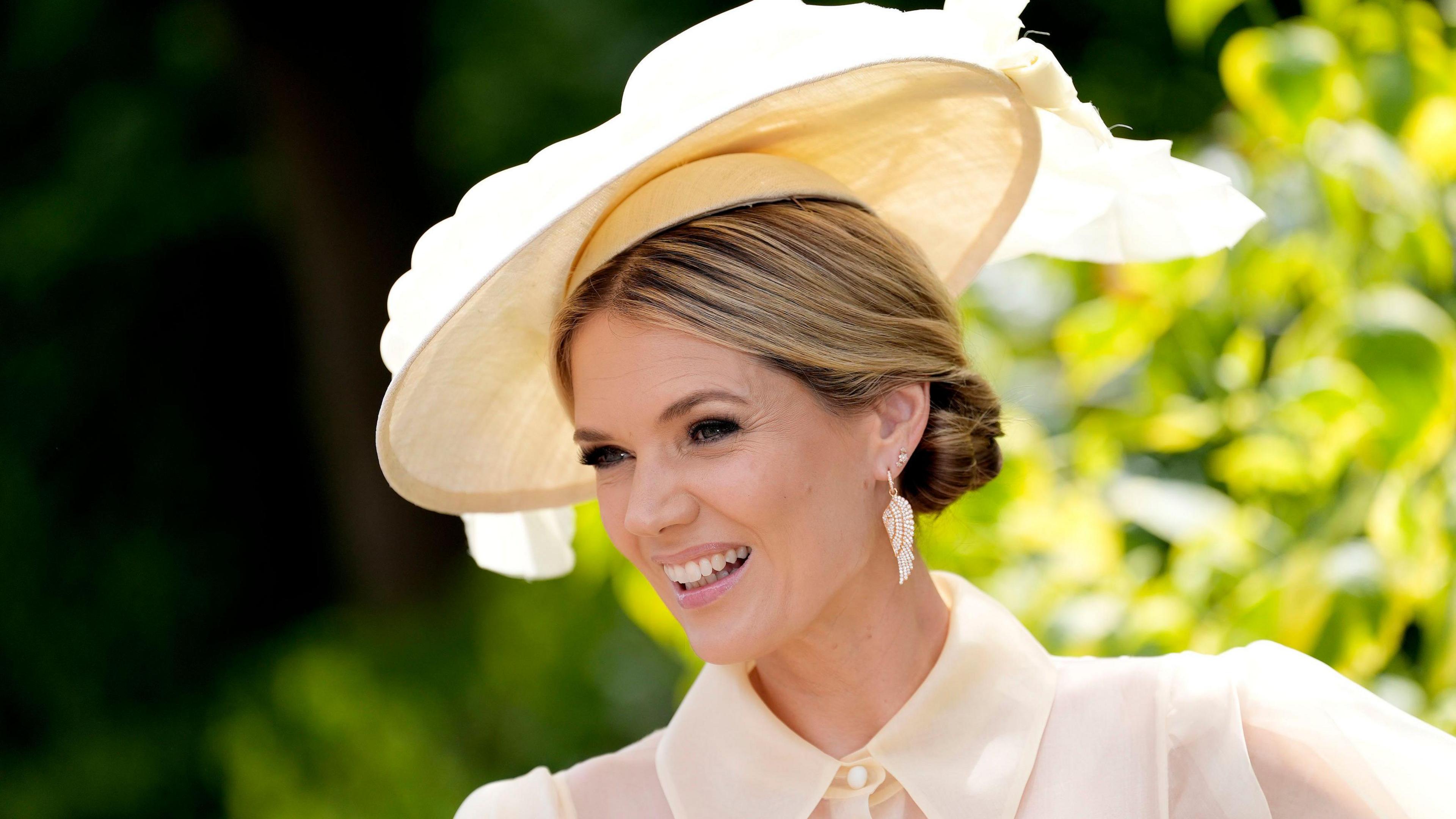 Charlotte Hawkins is smiling looking slightly off camera. The background is blurred but shows some greenery from plants. Charlotte has blonde hair which is tied back in a low bun hairstyle. She has a cream wide brimmed fascinator hat with large bow detailing on the top of it, large pearlescent angel wing earrings and a matching cream sheer blouse, which is buttoned up to the collar with a pearl button.