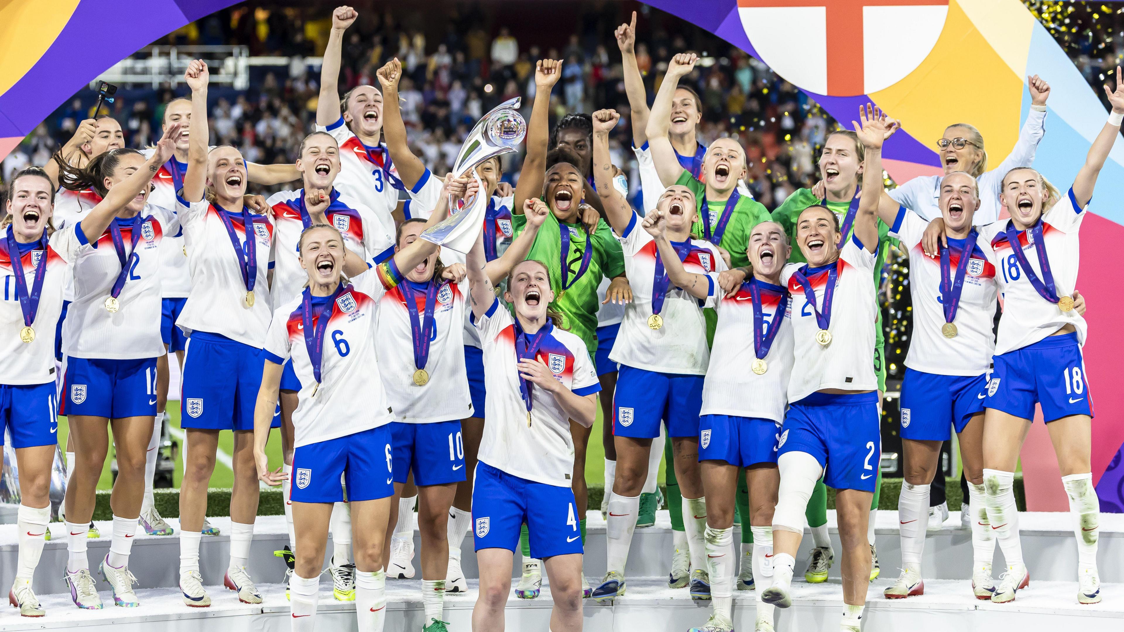 The Lionesses team stand on the podium, each player wearing the England jerseys and a gold medal with their fists in the air in celebration. 