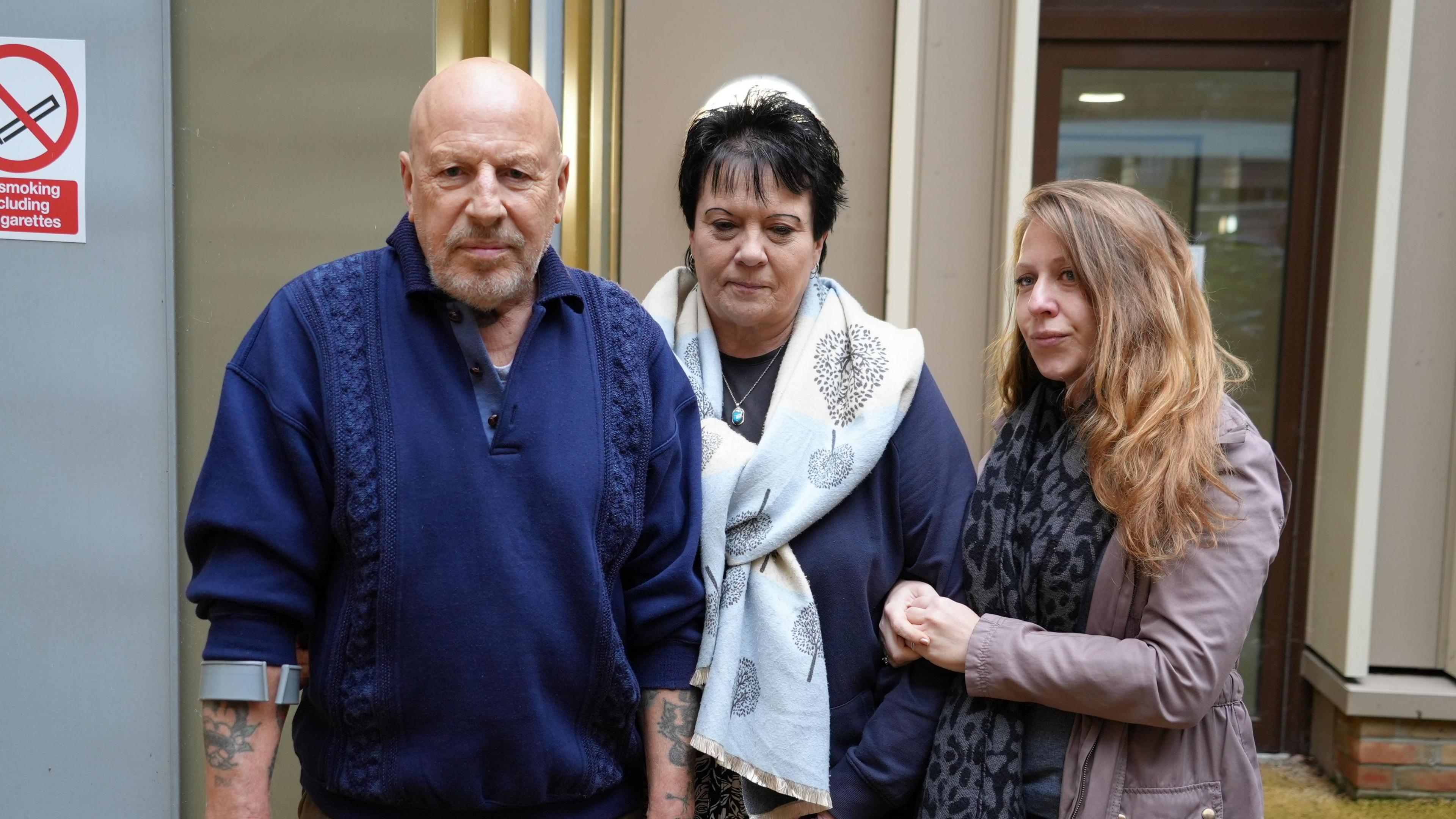 Holly Goodchild's family posing for a picture outside a court room at Norfolk Coroners' Court, including her dad - a bald man with tattoos and a crutch wearing a navy jumper - his partner, a woman with short black hair wearing a large white scarf over a navy top, and Holly's sister - a young woman with shoulder-length blonde hair, wearing a light purple jacket and a black and grey scarf.