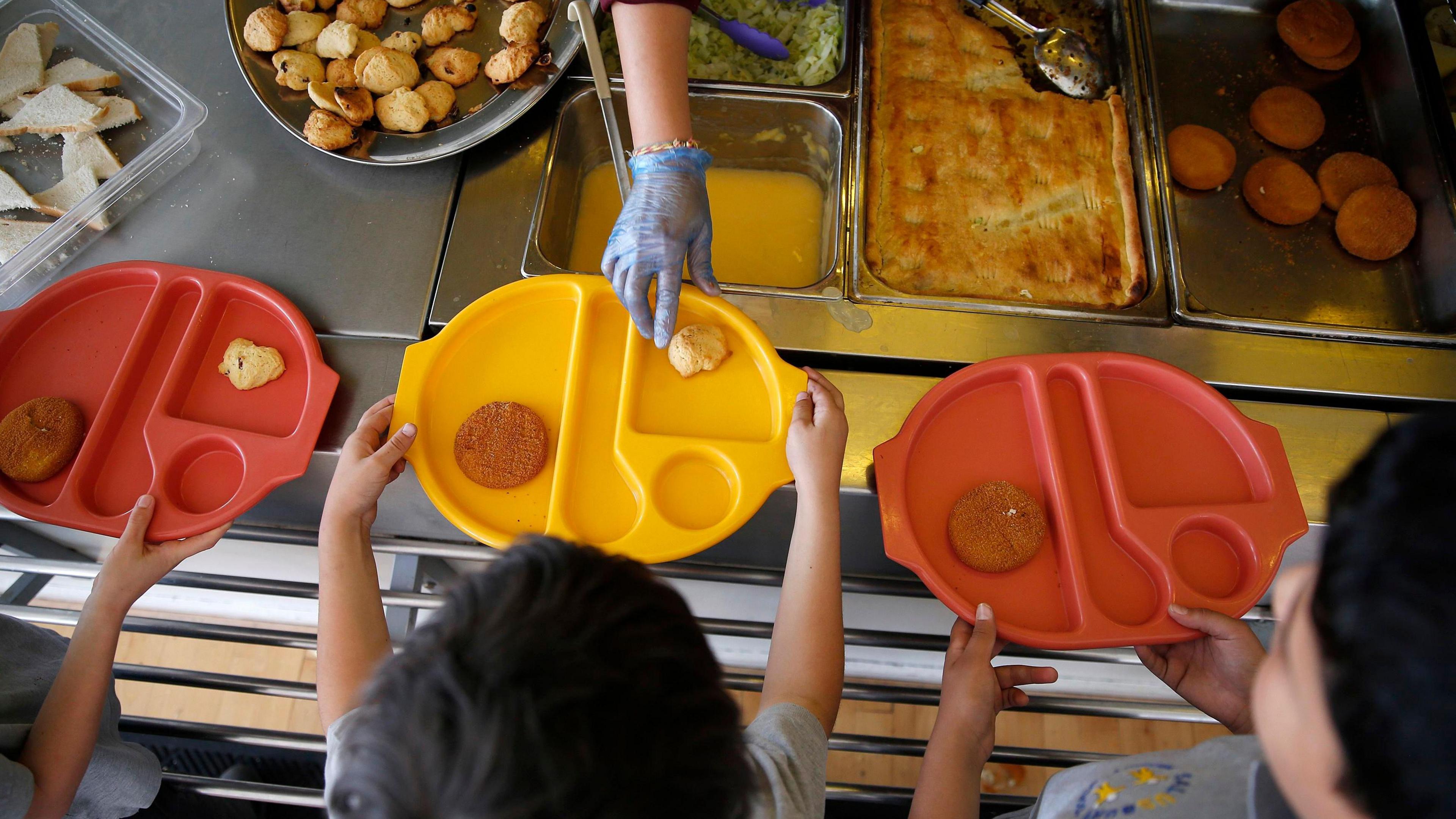 Three young children photographed from above hold out brightly coloured plastic trays with some food on them as more food is placed on one of the trays. In front of the children is an array of food, including roast potatoes and a pie in a metal tray. 