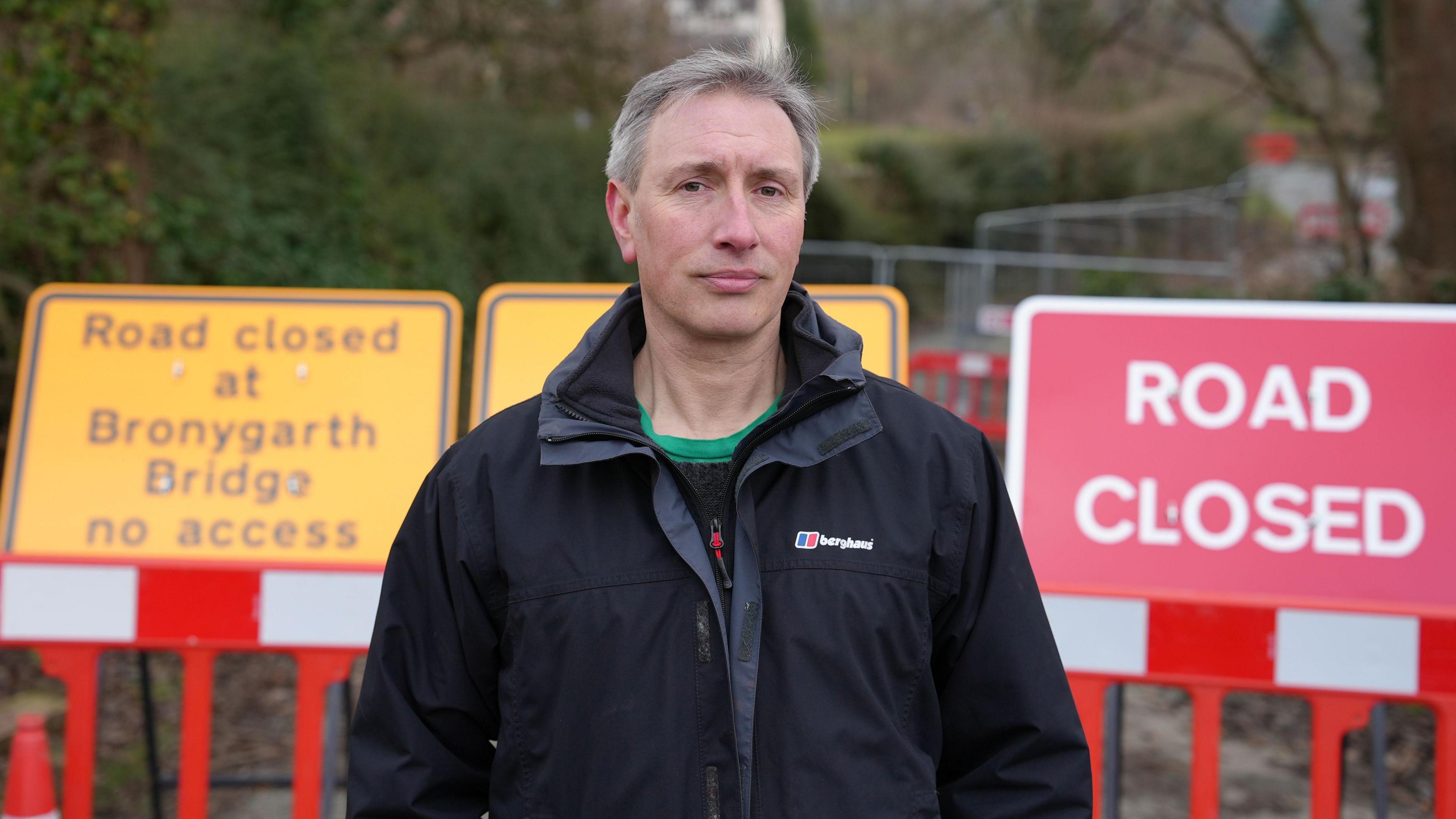 Paul Herbert, who has short grey hair, stands infront of yellow and red road signs which read "road closed at Bronygarth Bridge no access" and "road closed" He is wearing a black coat and black t-shirt with a green collar. 