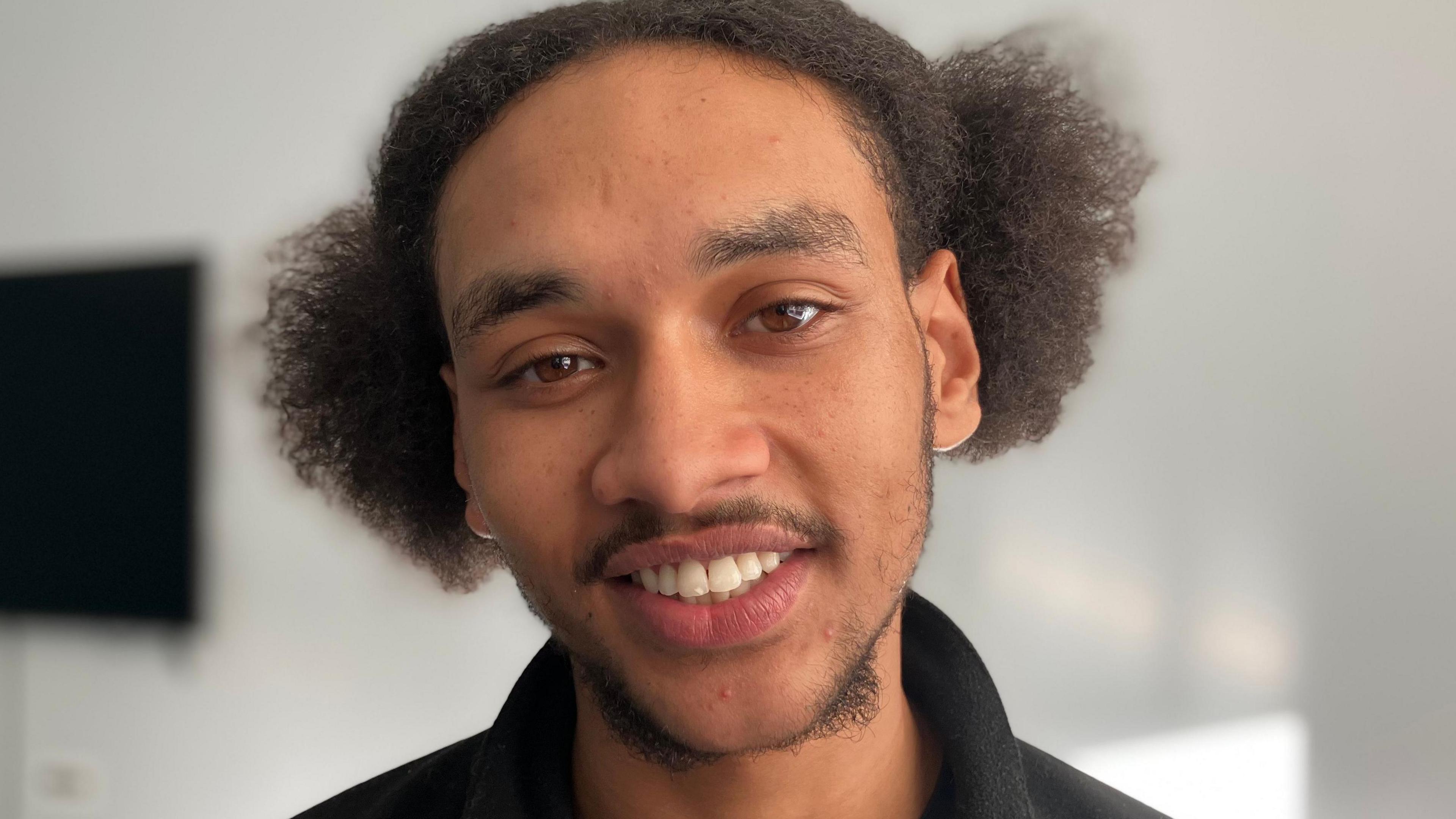 A young black man smiles at the camera in an empty classroom in a head and shoulders shot. He's wearing a  black top. 