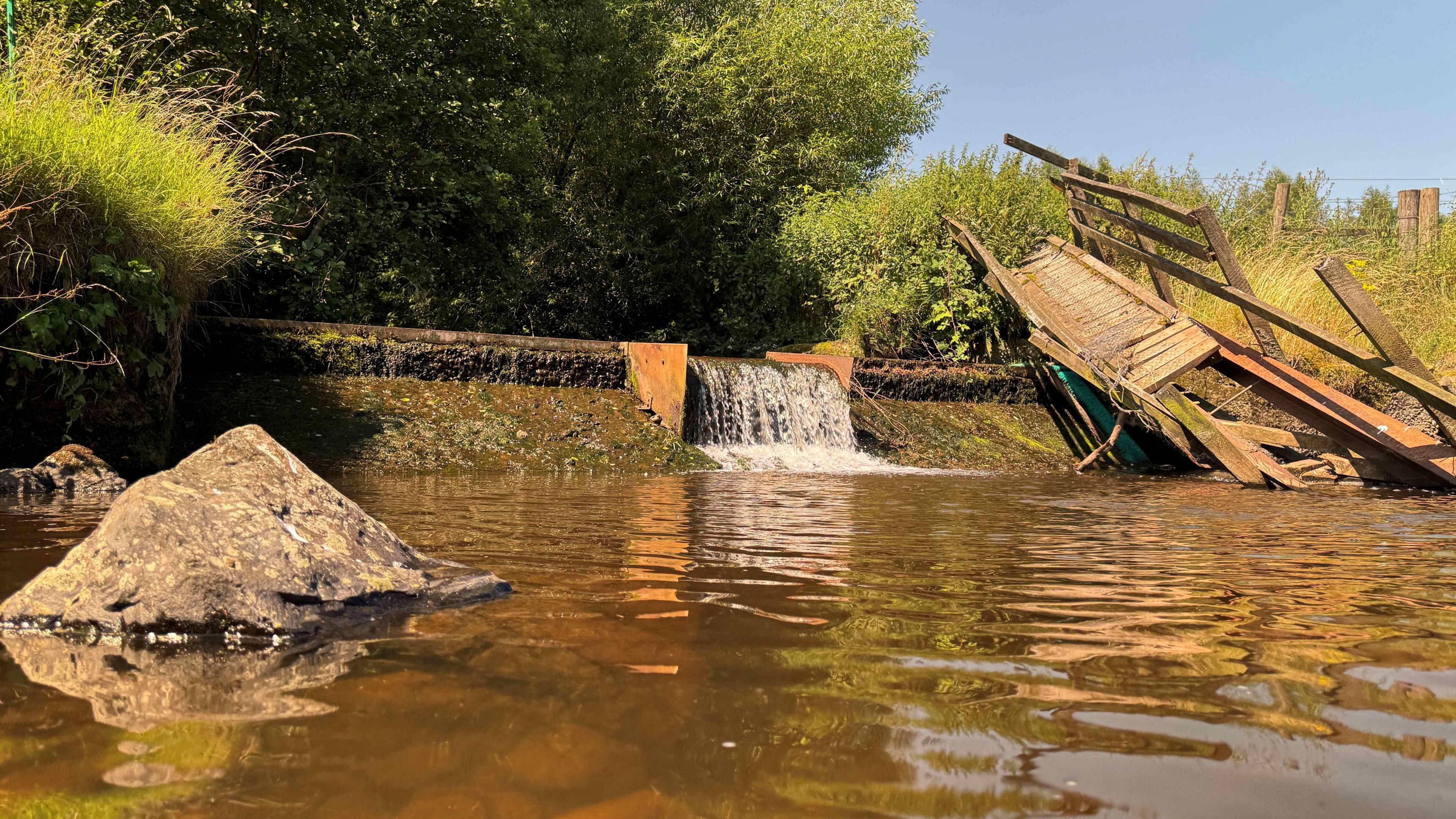 The redundant weir at Glinger Burn, with a pier-like structure falling into the river. The water is brown and the banks are grassy and green.