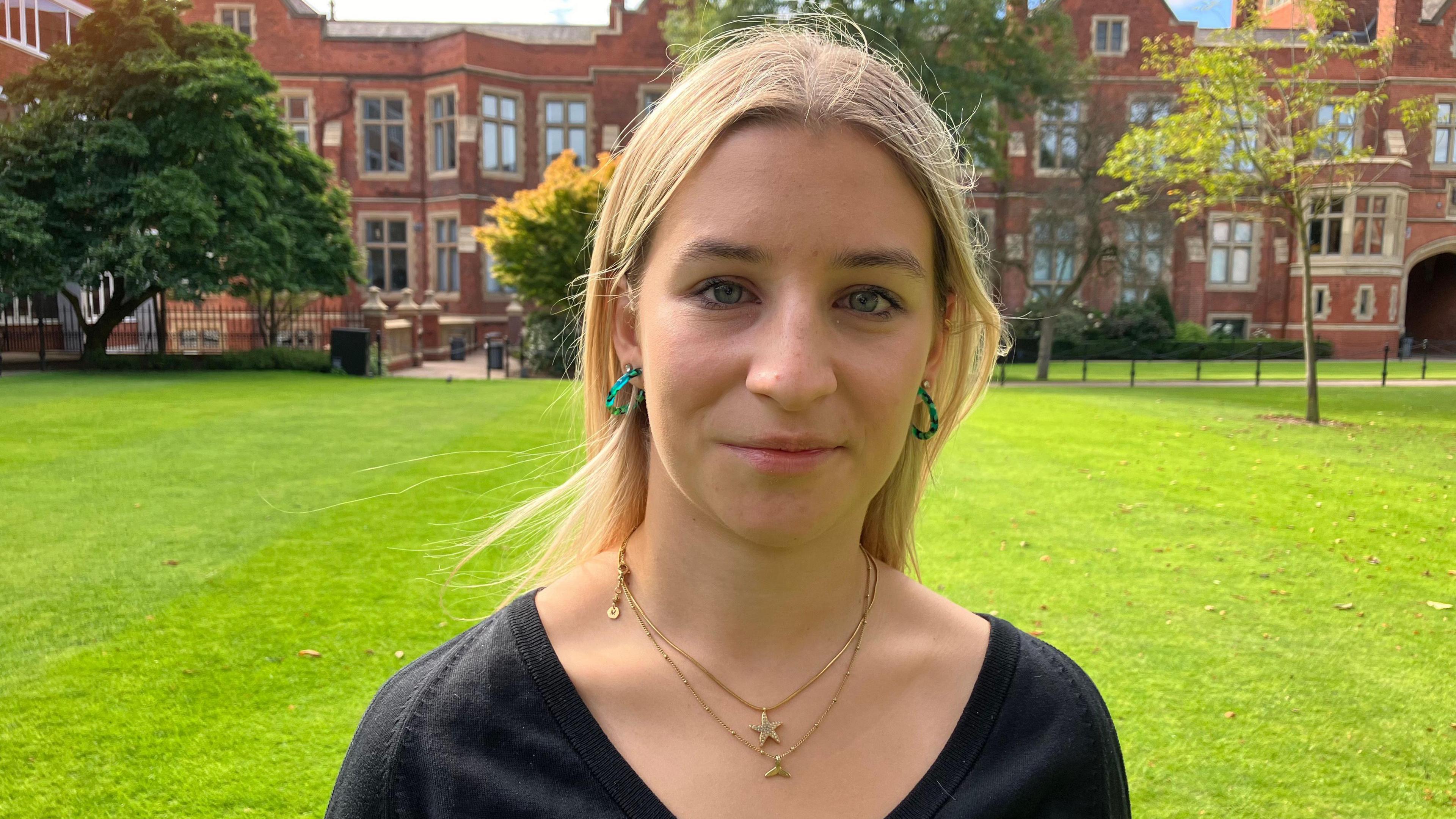 Lleucu is standing in front of the red bricks of Queen's on a grassy area. 

There are trees behind her and the grass is freshly mowed. 

She has blond hair in a middle parting, blue eyes, green hoop earrings and a double gold necklace on. 

She is wearing a black v neck top and is looking directly at the camera. 