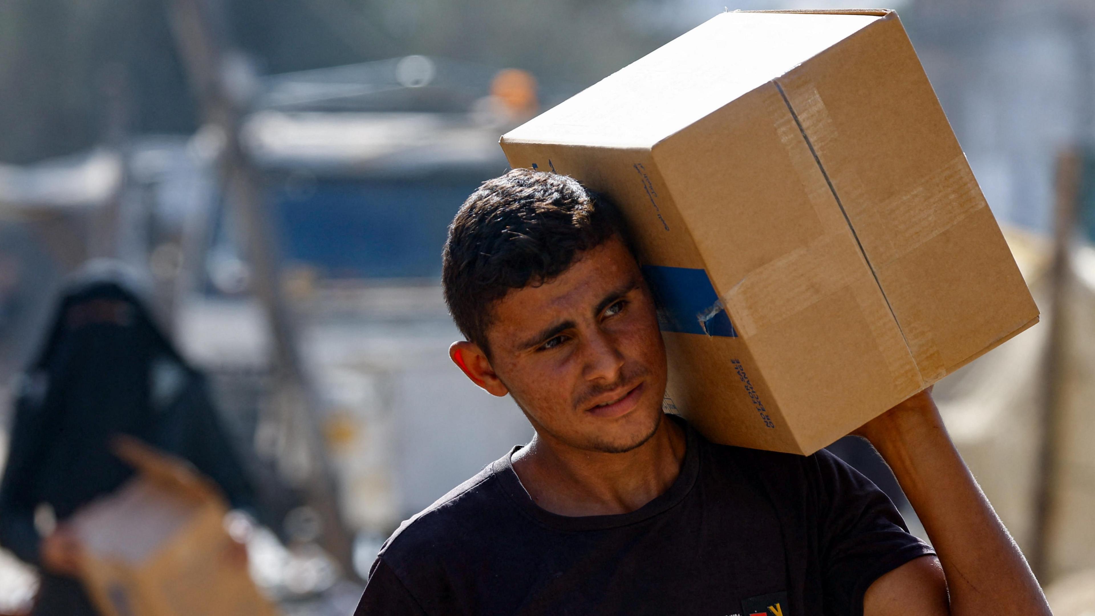 A man with short dark hair carries a cardboard box of aid on his shoulder - a woman carrying a box can be seen in the background behind him