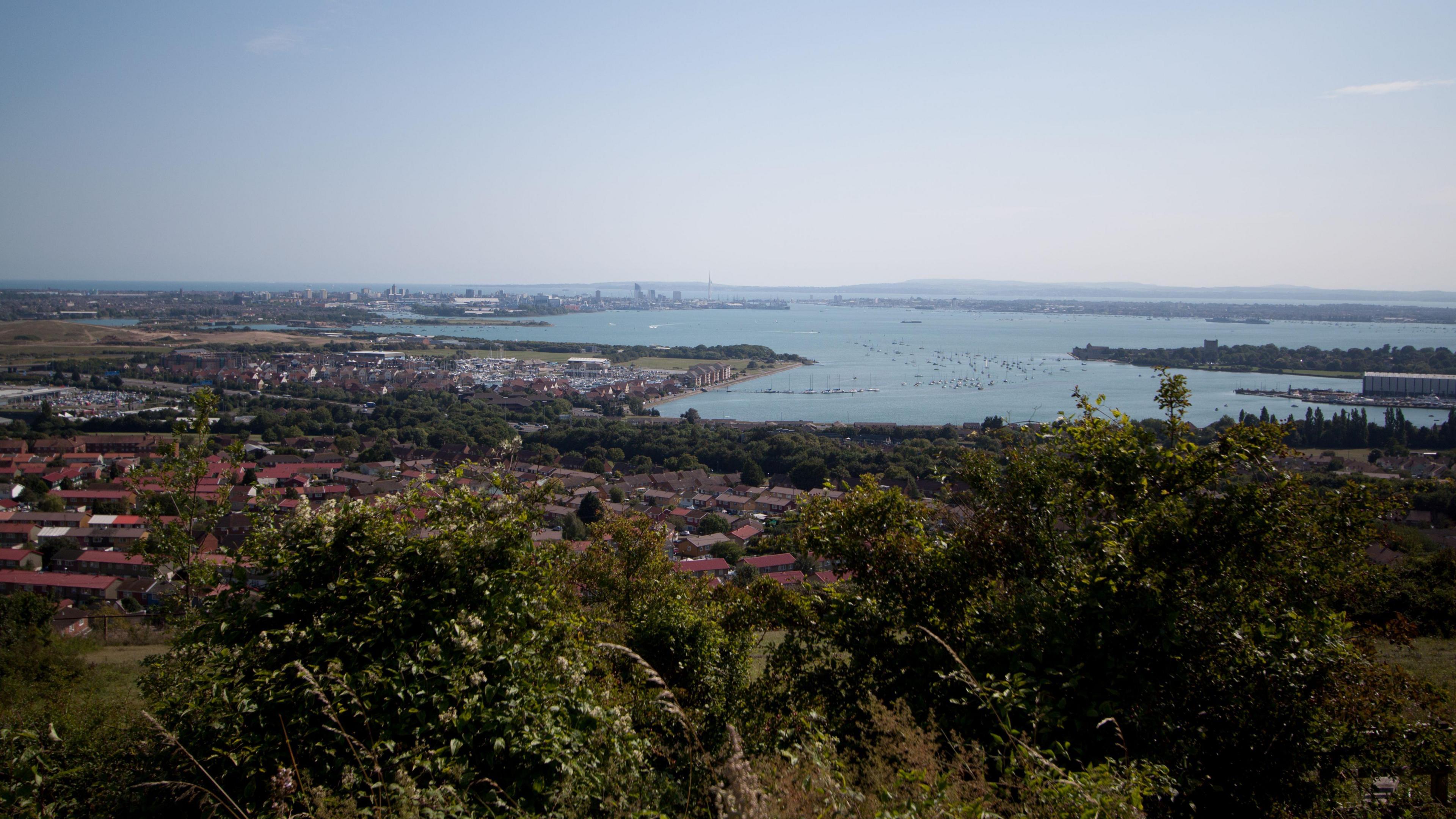 The view from Portsdown Hill, which overlooks Portsmouth there is trees and other greenery in the foreground and housing and the sea in the background