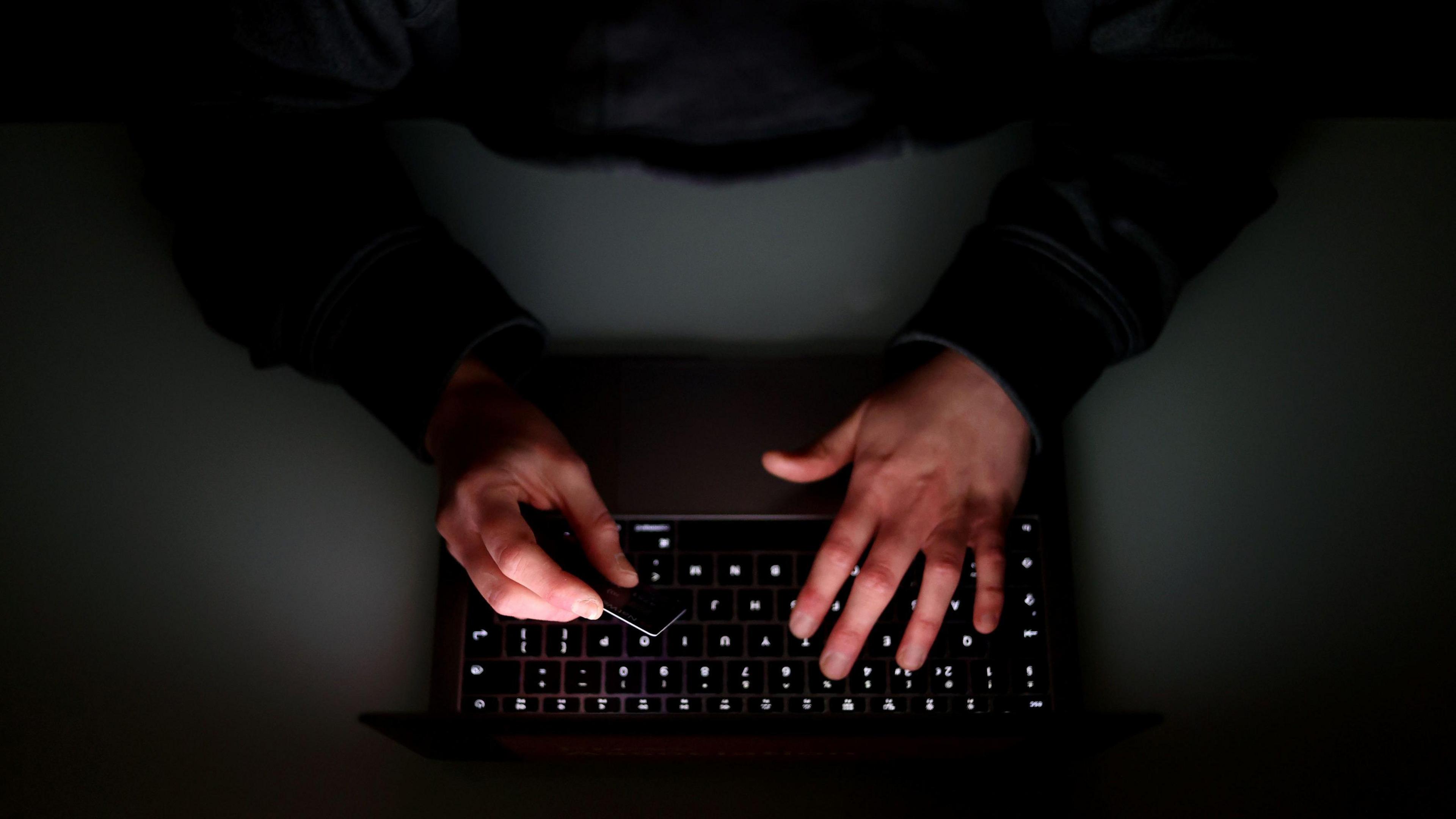 A laptop user with his hood up holding a bank card. The user is dressed in a black long-sleave hooded top and the laptop keypad is also black