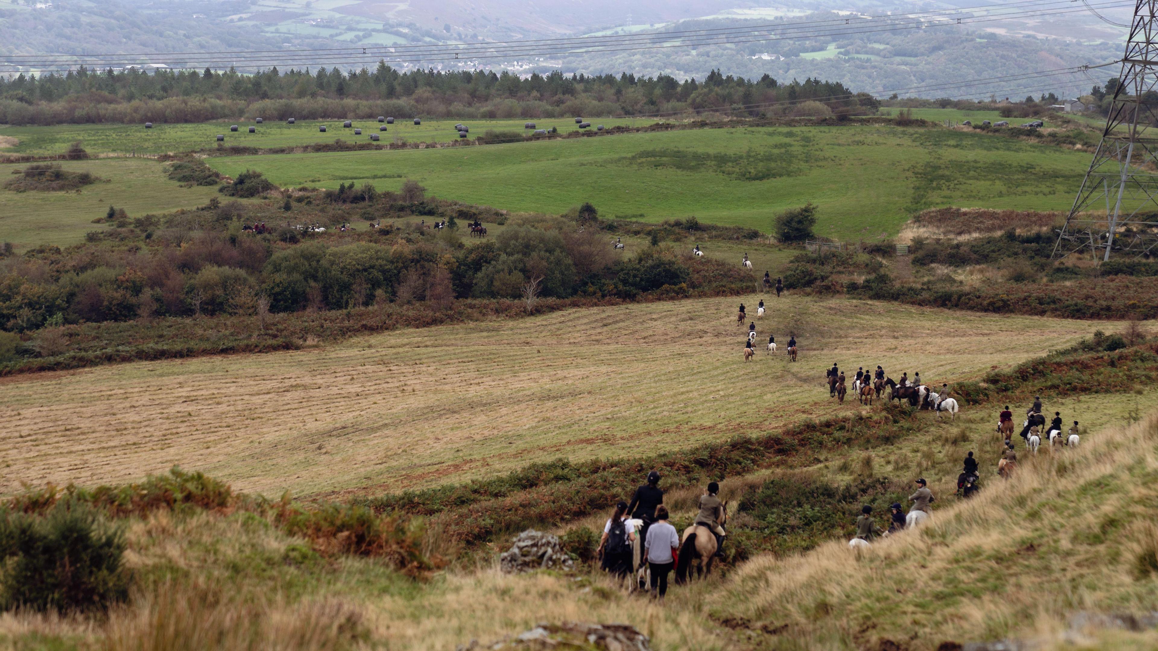 Mae gorymdaith o deuluoedd ar geffylau yn dilyn lawr allt. Yn y llun mae modd gwerdd nifer o gaeau gwyrdd.