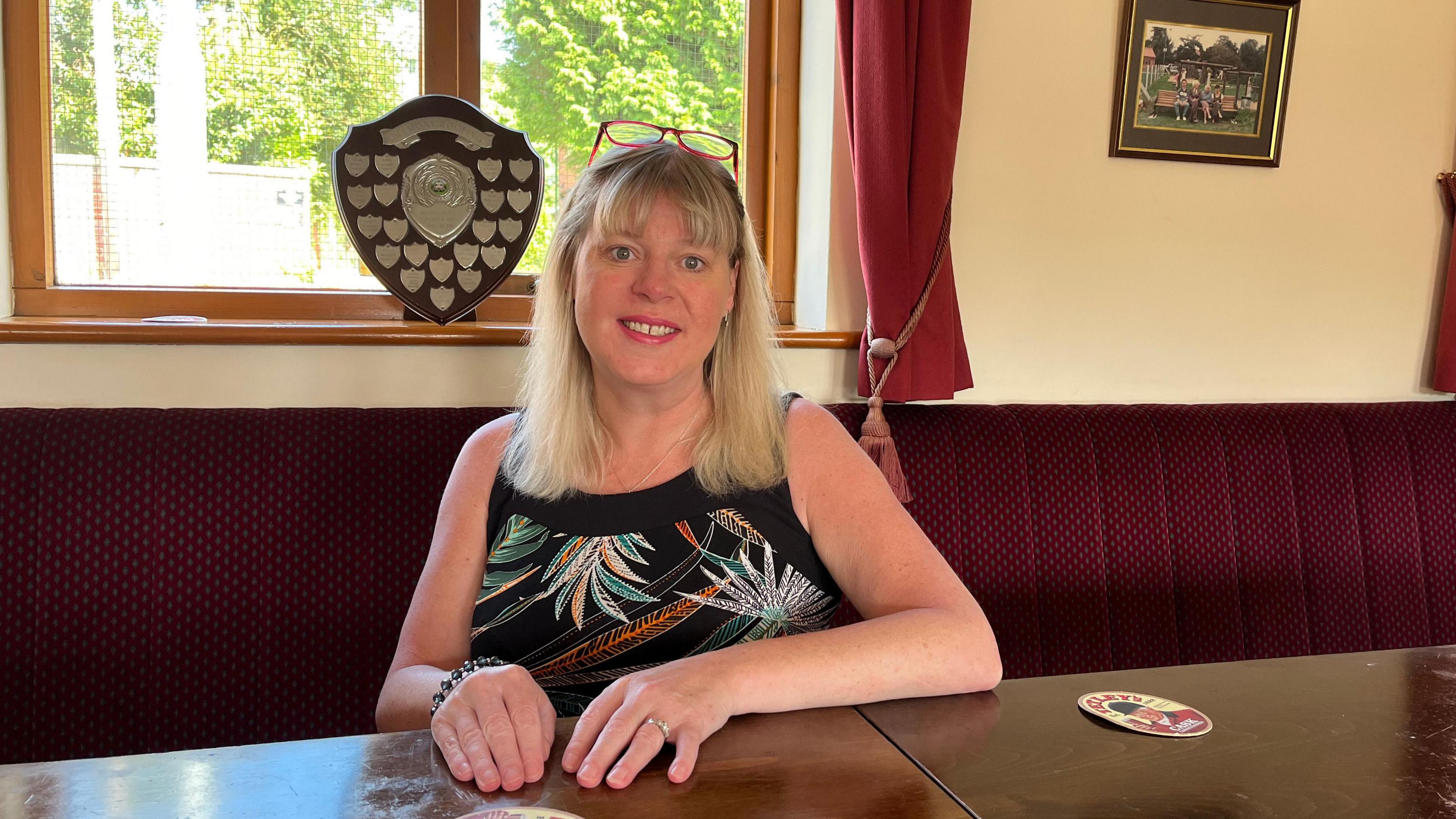 Jane Naylor, Paddy's daughter, sits in the club house on a red-cushioned bar bench. She is blonde and wearing a black vest top, with embroidered leaves on it. Behind her is a shield-shaped trophy won by the local football club. 