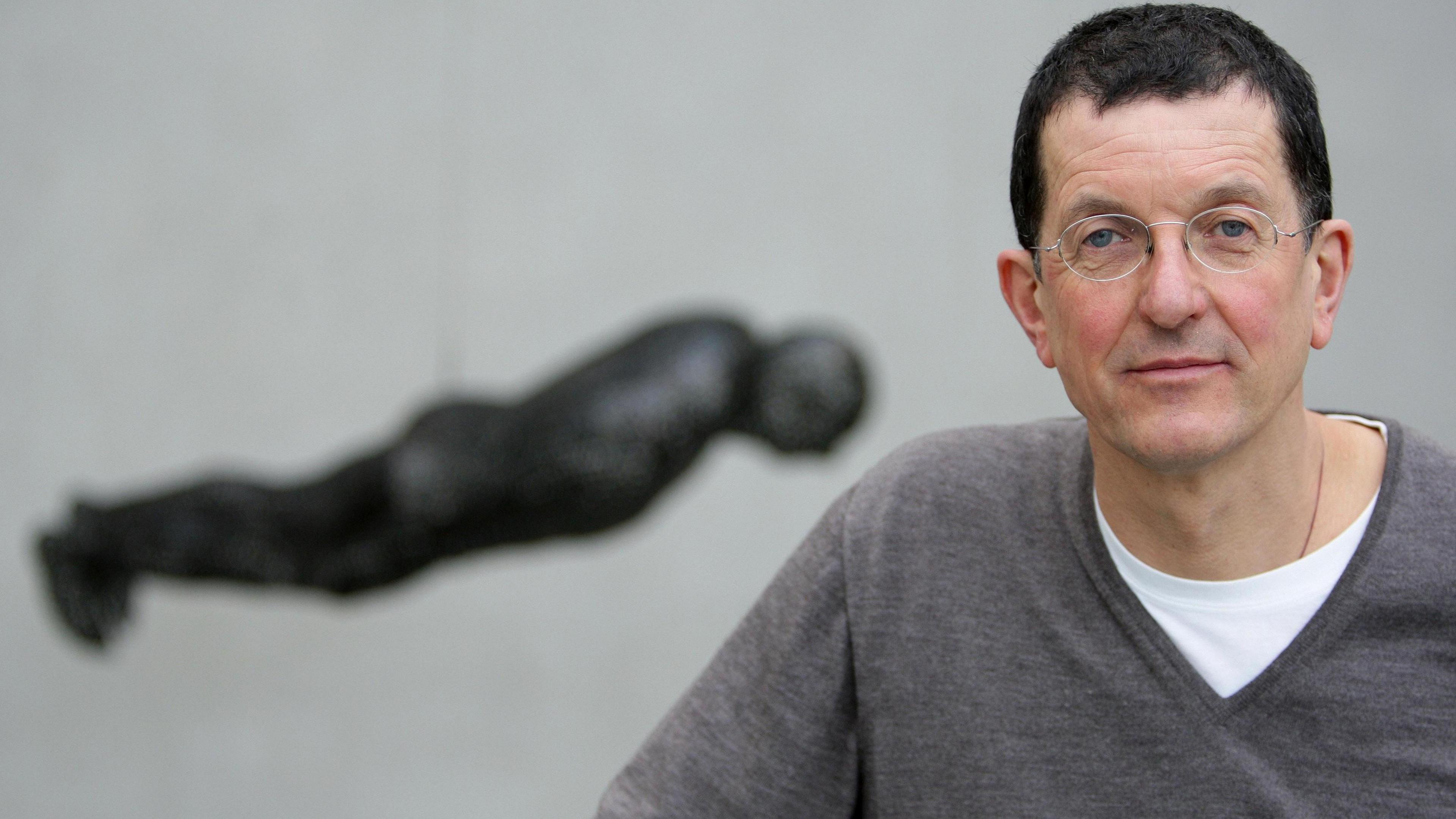 Sir Antony Gormley, who has short black hair and rounded spectacles, poses for a picture with a sculpture based upon his own body hanging in the background.