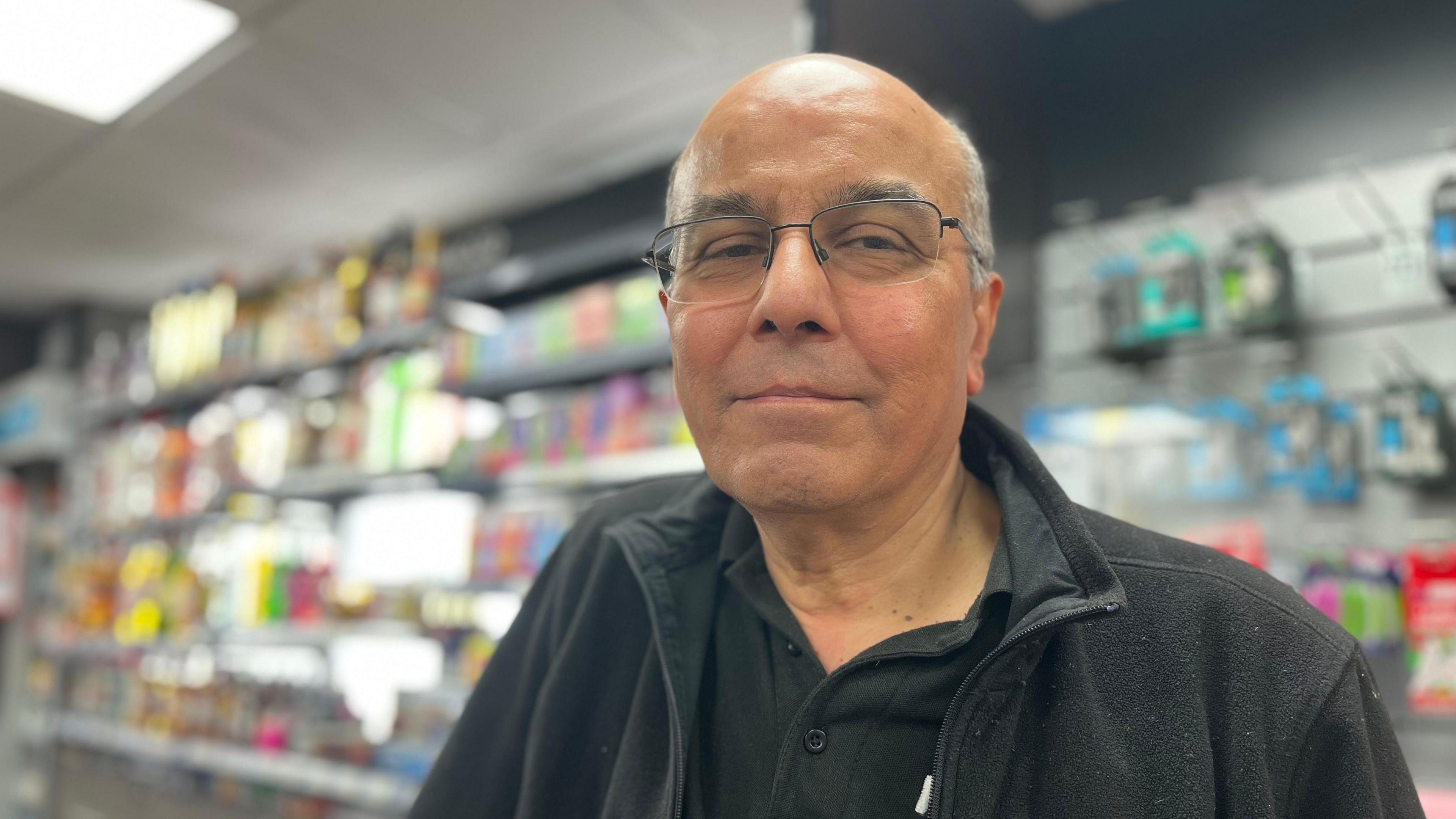 Vidyut Soni looks directly at the camera, unsmiling. He is wearing glasses and a black polo shirt and fleece. Behind him are shelves stocked with goods. 