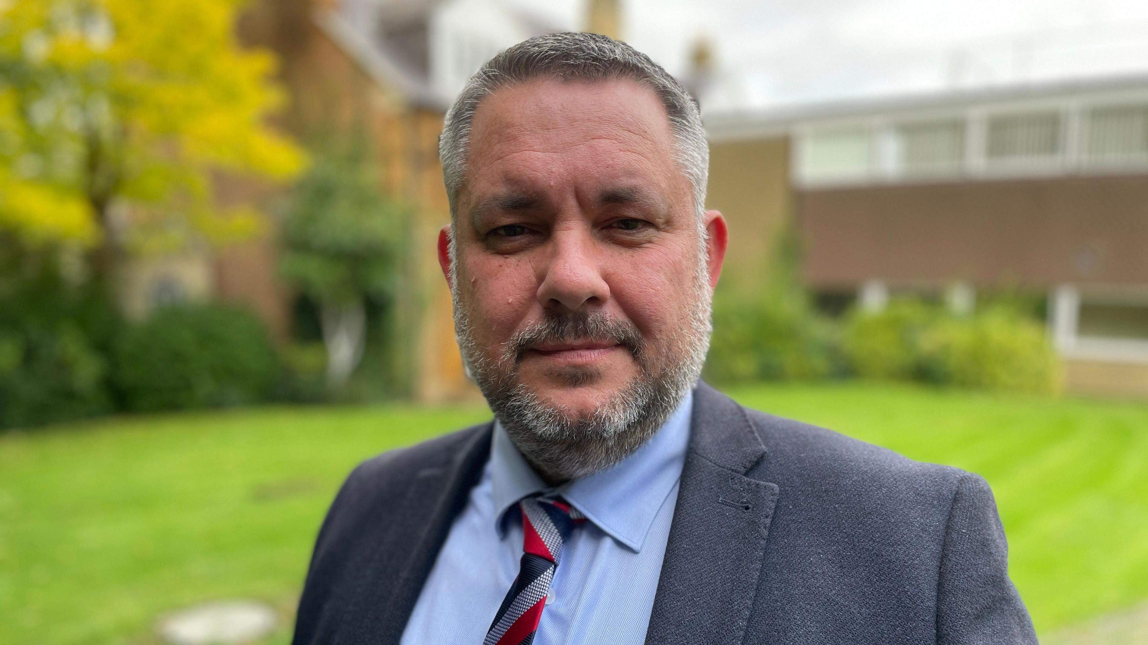 A man in a navy blazer, light blue shirt and red, grey and blue tie stands in a garden looking directly at the camera