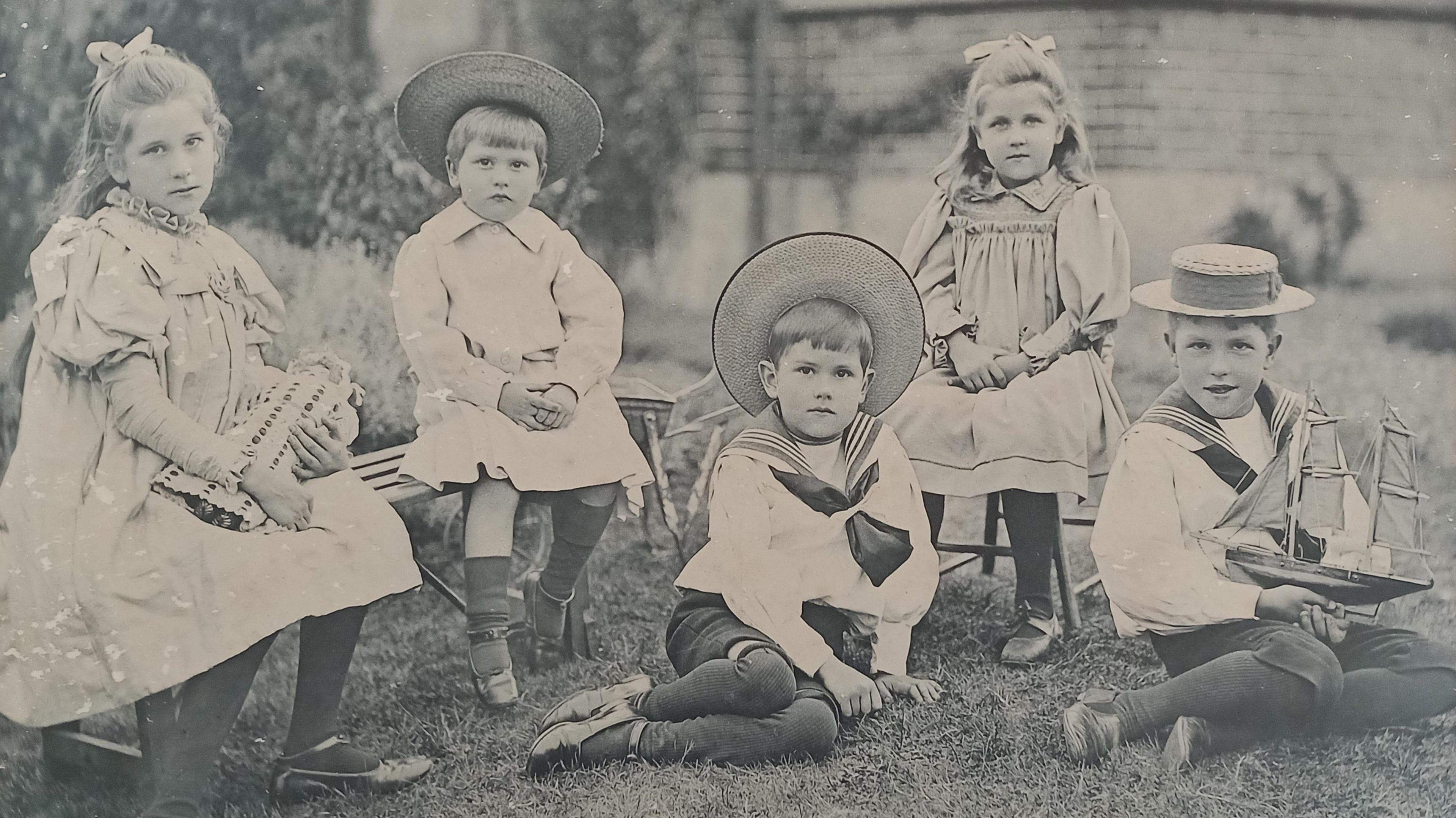 A black and white image of Noel Carrington and his family. There are five children, all sat down, with two sat on the grass, one holding a boat. Three have straw hats on and two girls are sitting down, with bows in their hair. The girl on the left is holding a doll. They are in front of a building.