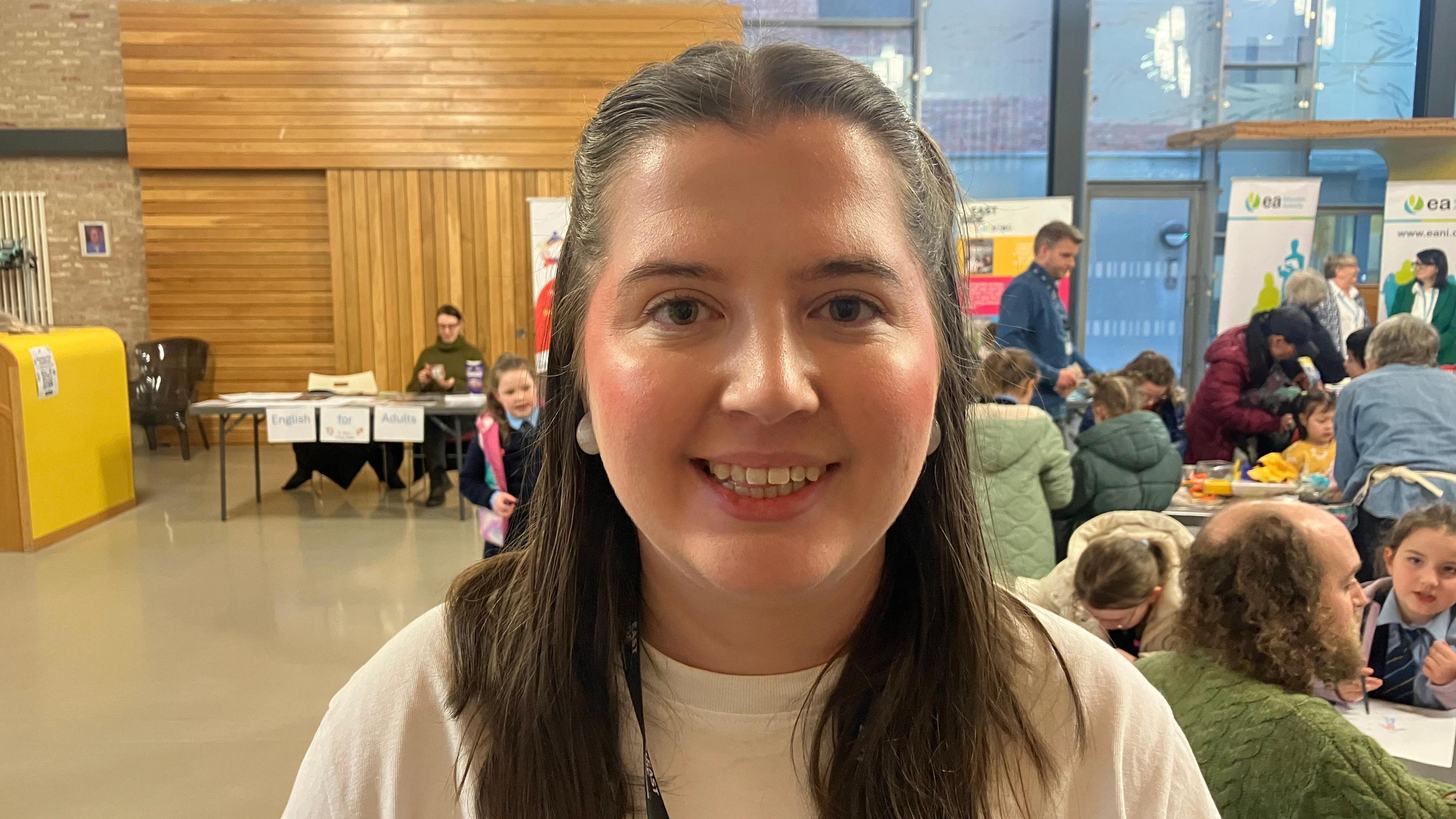 A woman with brown hair wearing a white t-shirt looking at the camera. Behind her there are kids and adults sitting at tables 