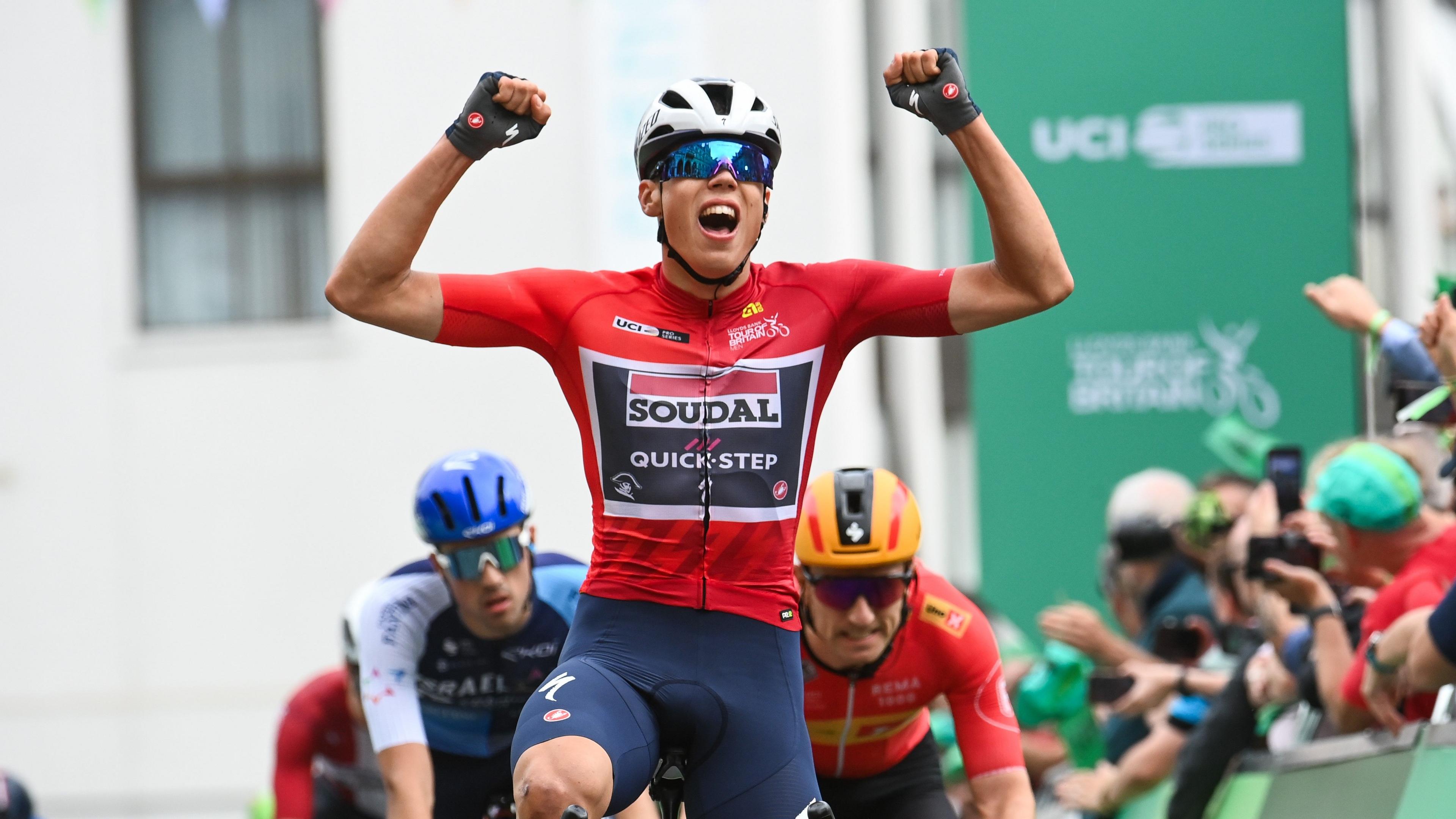 A cyclist with his arms aloft as he crosses the line first. He is wearing a red top and a cycling helmet and wrap around sunglasses. He is being followed by other cyclists and there is a crowd of people on the right hand side behind the hoardings.